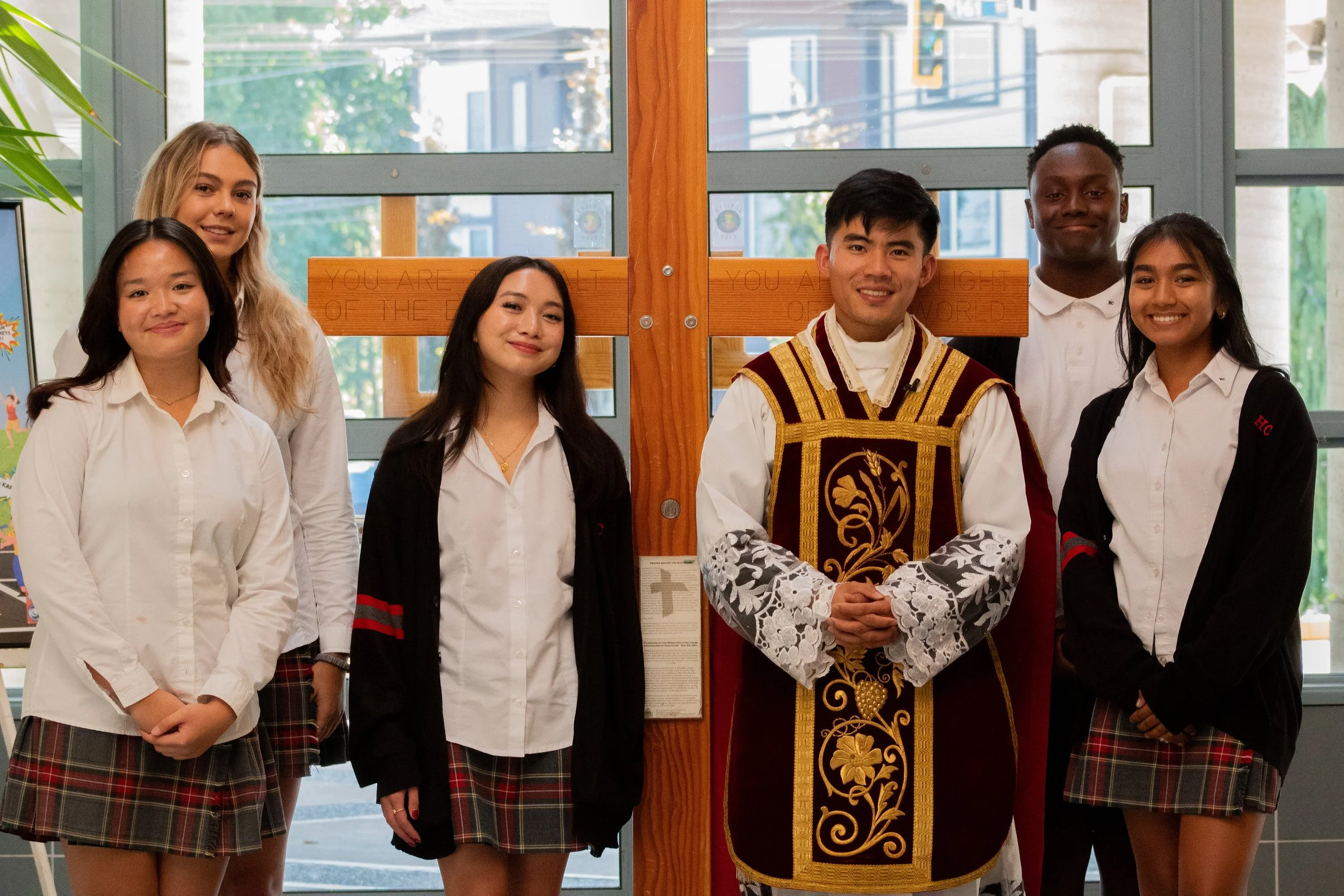 Group of six students standing inside school, posing with a priest dressed in ornate vestments. The students are wearing white shirts and plaid skirts or pants. Behind them, large windows reveal the outdoors.
