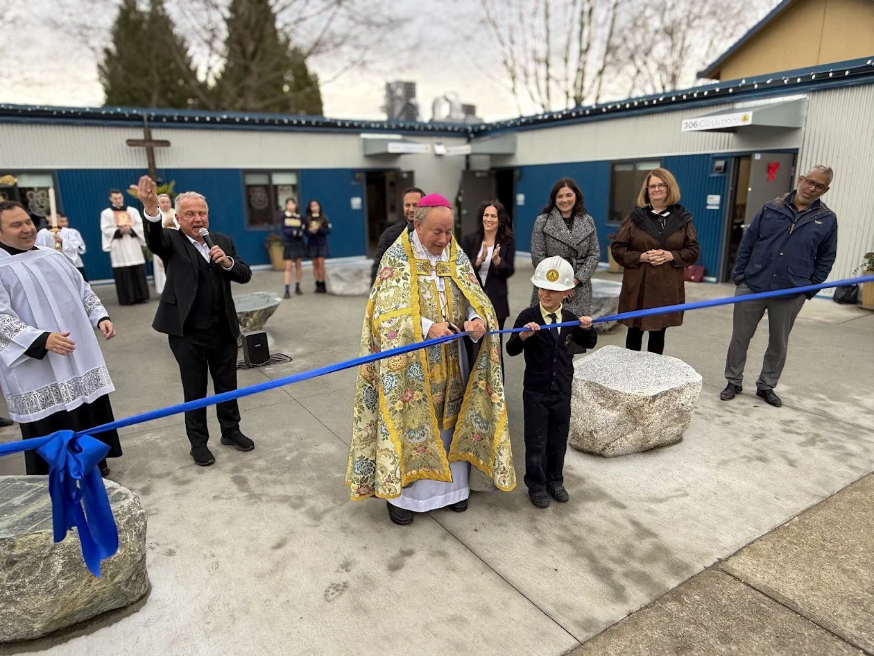 A ribbon-cutting ceremony with a religious figure in ornate vestments and a bishop's mitre, a young boy in a helmet, and several onlookers, outside a building with string lights and a cross on the wall.