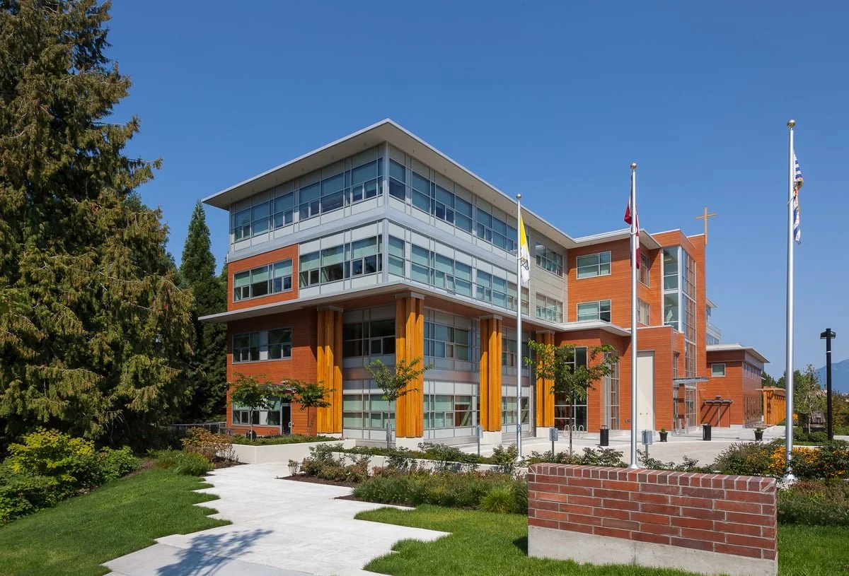 Modern four-story building with brick and glass facade, featuring multiple flagpoles and a landscaped garden, set against a clear blue sky.
