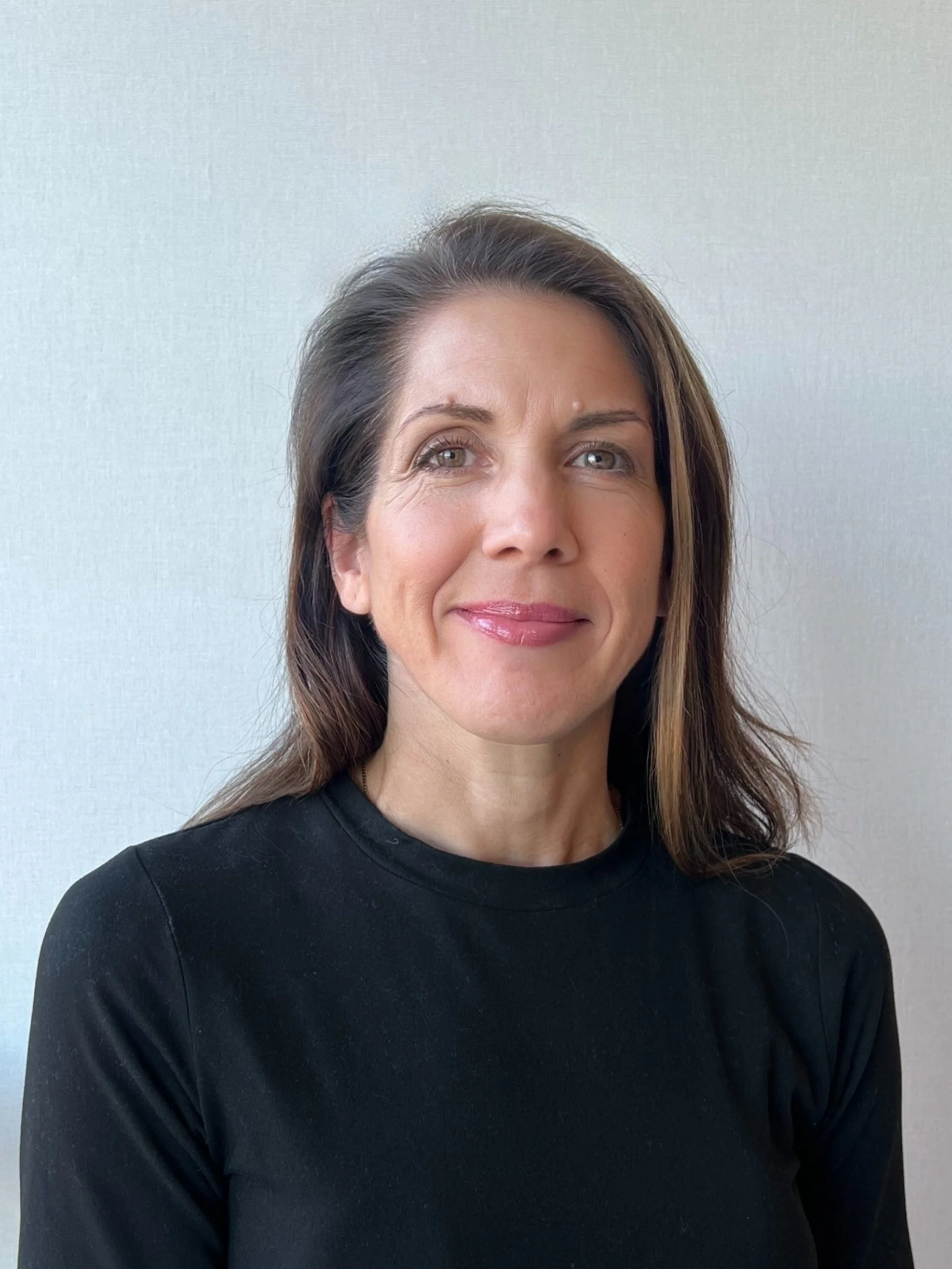 A woman with shoulder-length brown hair, wearing a black top, smiling softly against a plain light background.