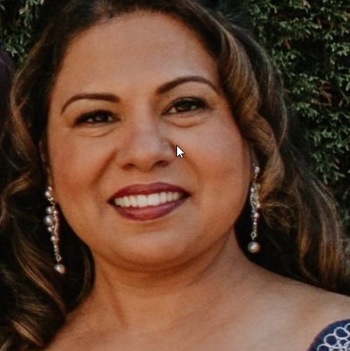Close-up of a smiling woman with wavy brown hair, wearing pearl earrings and a blue dress, outdoors with greenery in the background.