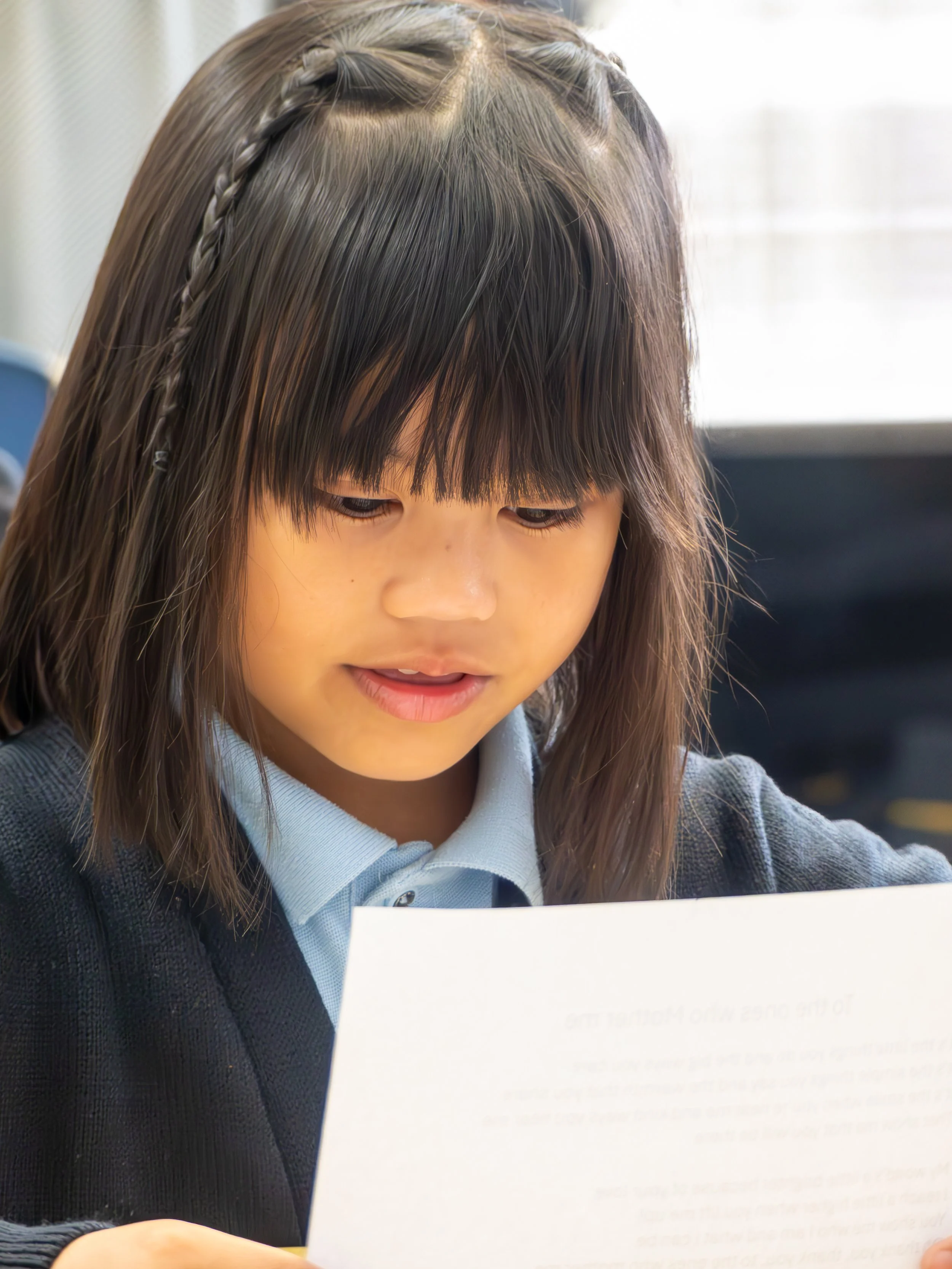 A young girl with dark hair and bangs, wearing a school uniform, looking at a piece of paper with a focused expression.