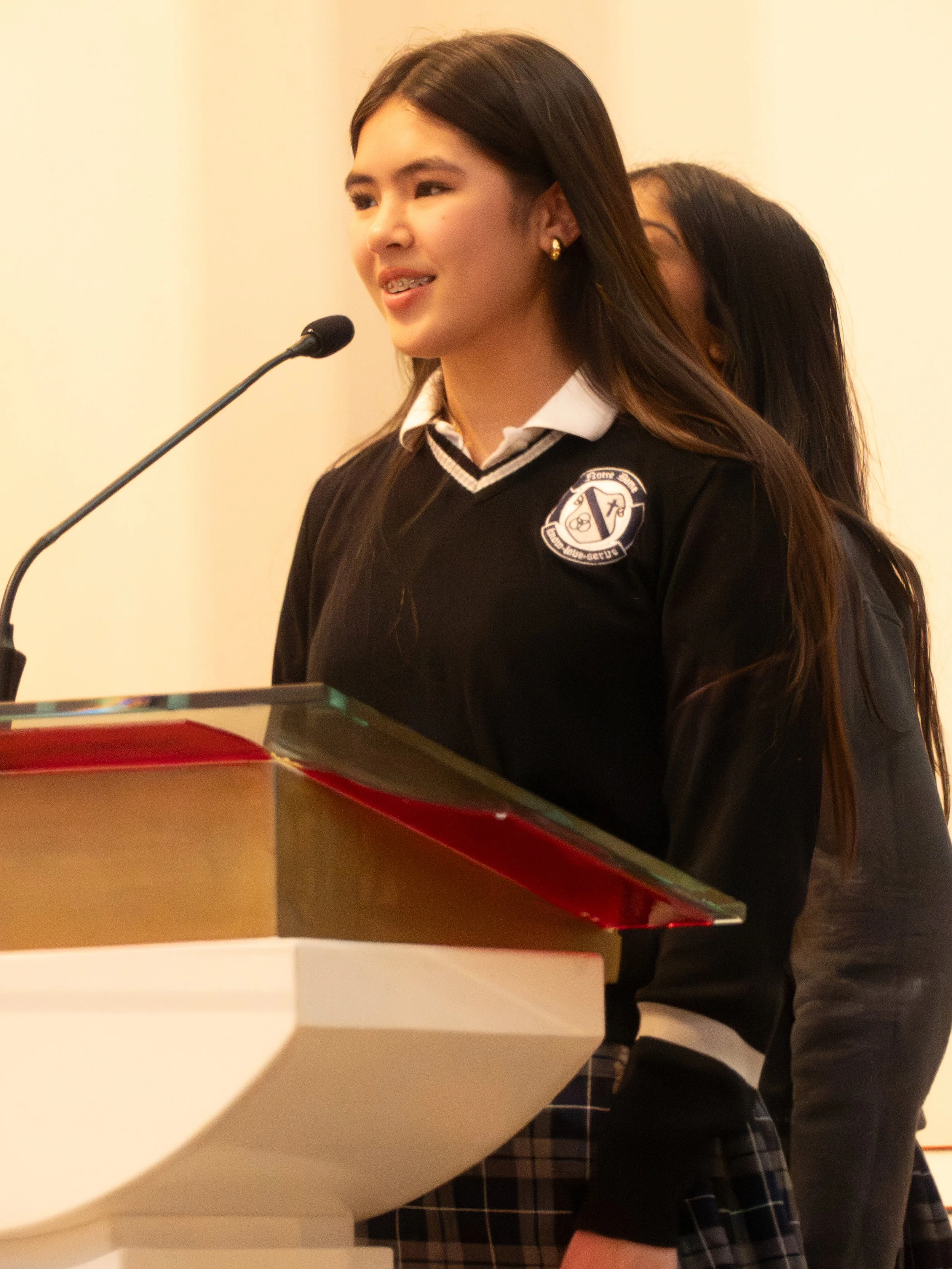 A young woman with long dark hair, wearing a black sweater, speaking at a podium with a microphone.