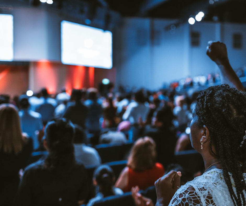 Crowd of people attending a conference or presentation in a large auditorium with a raised fist in the air.