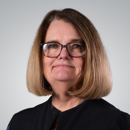 A middle-aged woman with shoulder-length light brown hair, wearing glasses and a black top, looking directly at the camera with a neutral expression.