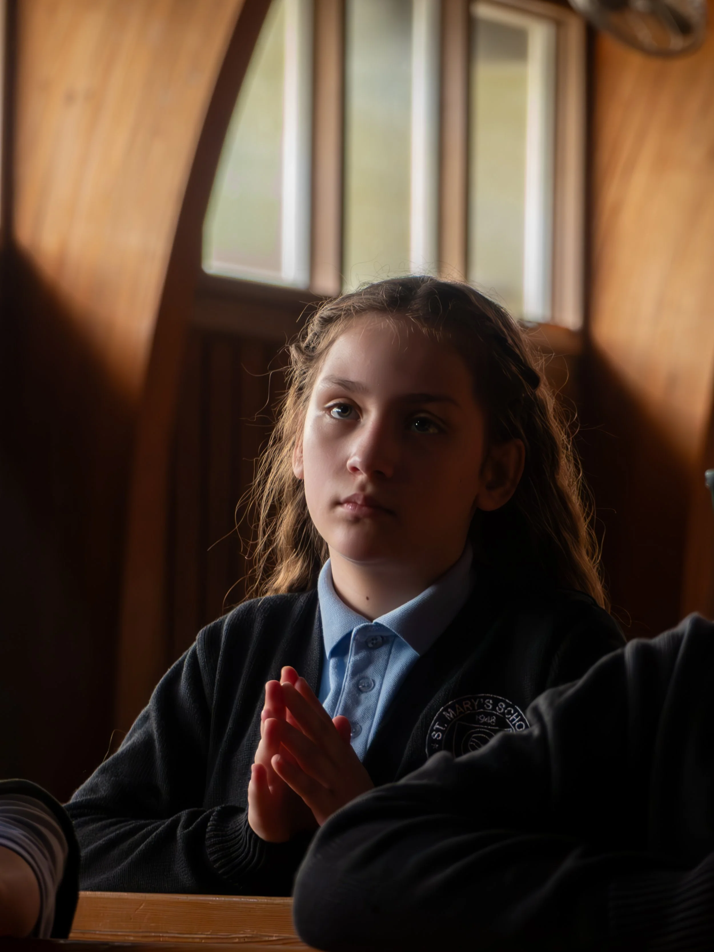 Young girl in school uniform sitting at a desk inside a room with wooden walls and windows, with her hands clasped together.