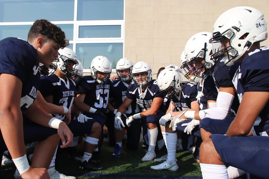 Young football players in navy jerseys and white helmets are gathered in a huddle outside a building during daytime.