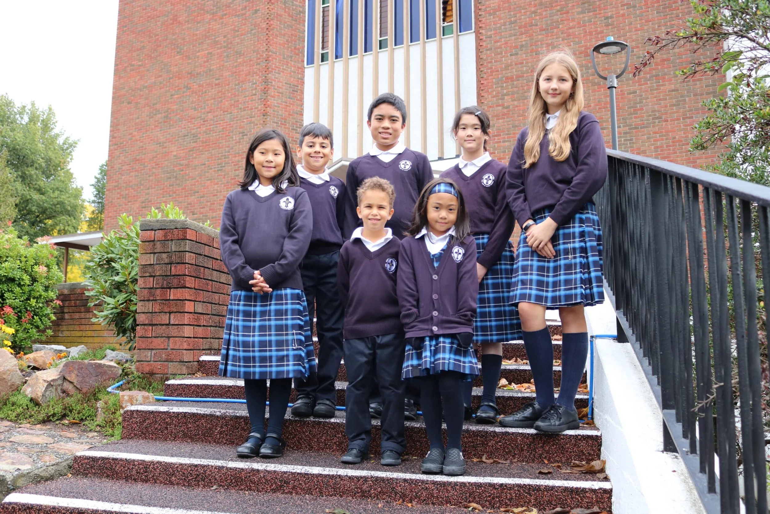 A group of school children wearing uniforms with plaid skirts and sweaters, standing on outdoor stairs with a brick building in the background.