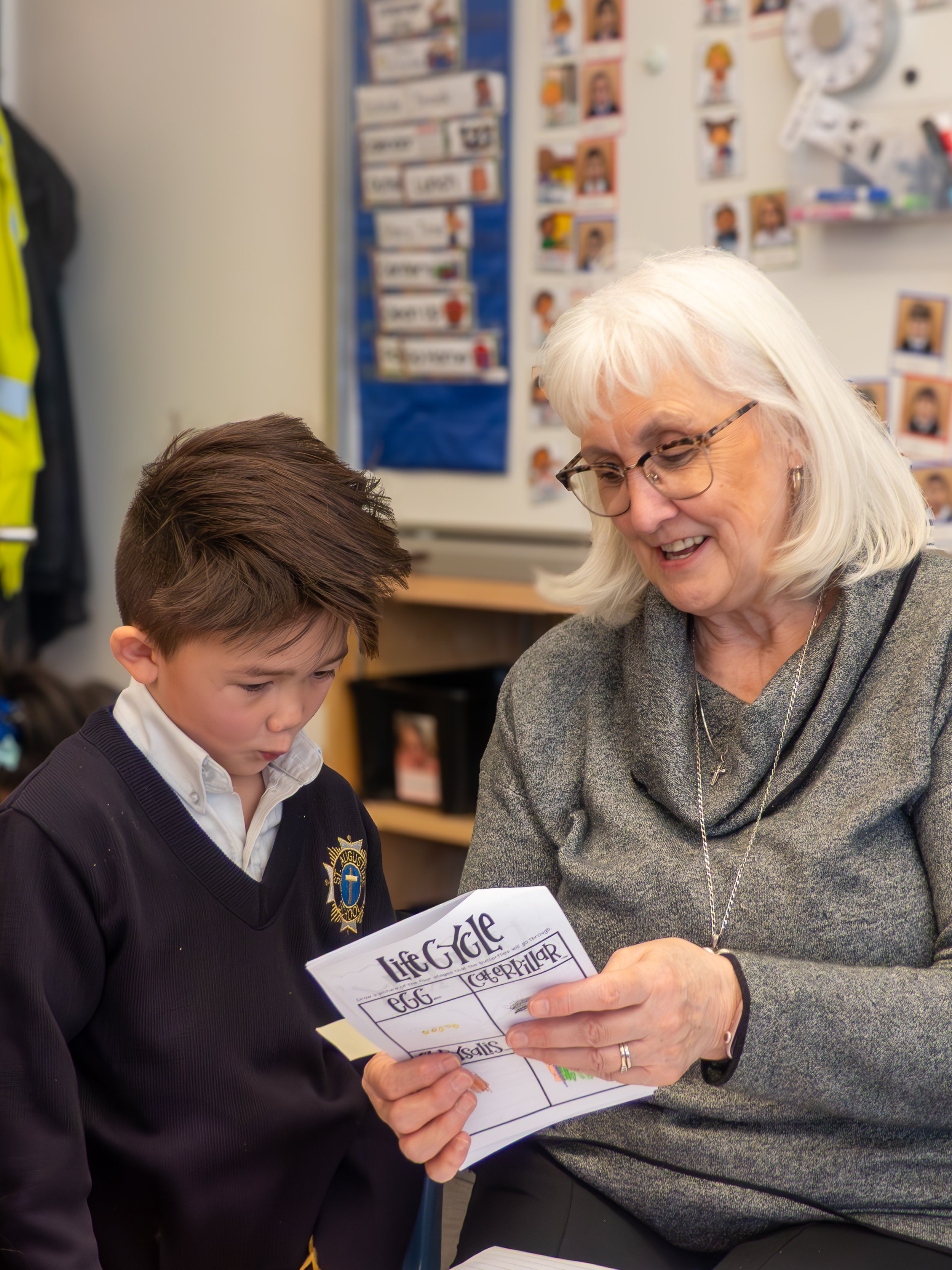 A young boy and an older woman, possibly a teacher, sitting together in a classroom. The woman is smiling and showing him a handwritten story or worksheet titled 'Life Cycle.' The classroom background includes a blue chart and photographs of children on the wall.