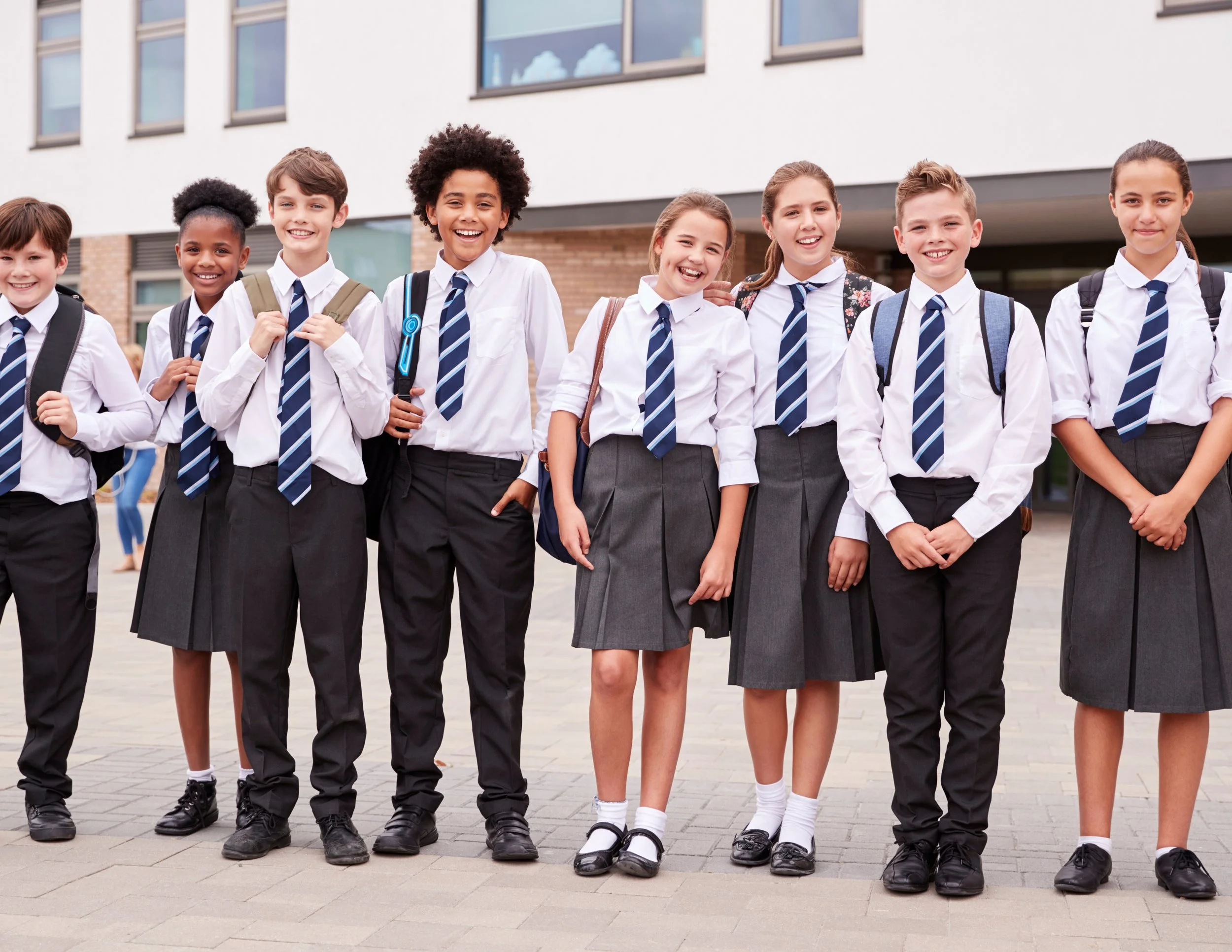 A group of eight diverse school children in uniform standing outside a modern school building, smiling at the camera.