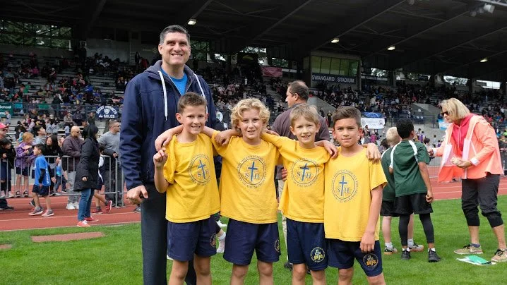 A coach with four young boys in yellow shirts and navy shorts on a sports field during a track and field event, with a crowd in the stands.