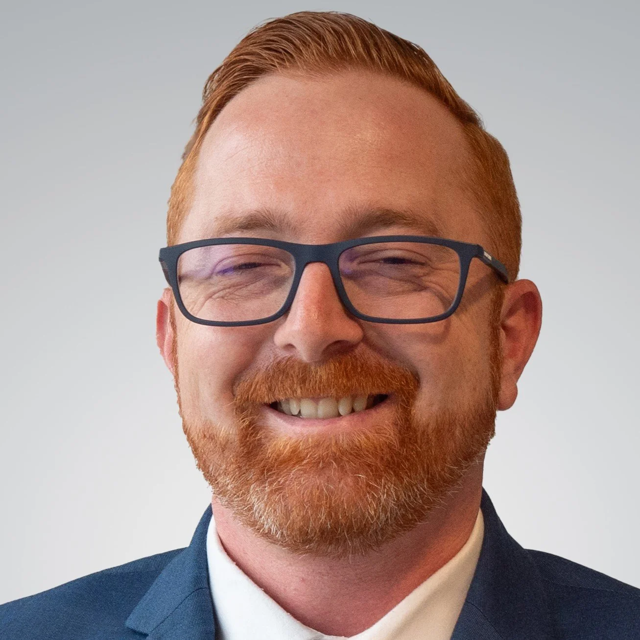 Close-up of a smiling man with red hair, beard, and glasses, dressed in a suit against a plain background.