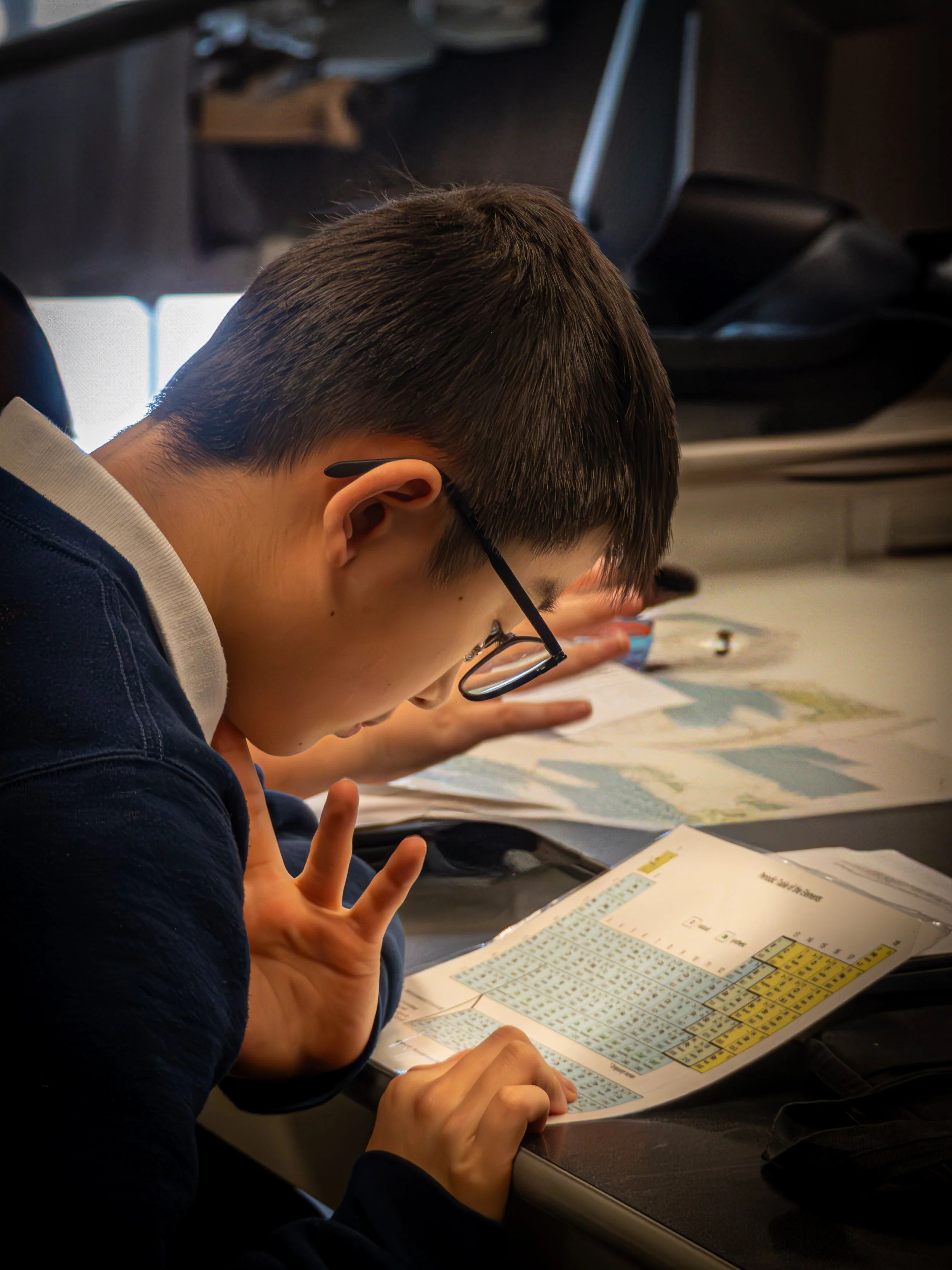 A young boy wearing glasses studies a periodic table with a peace sign gesture.