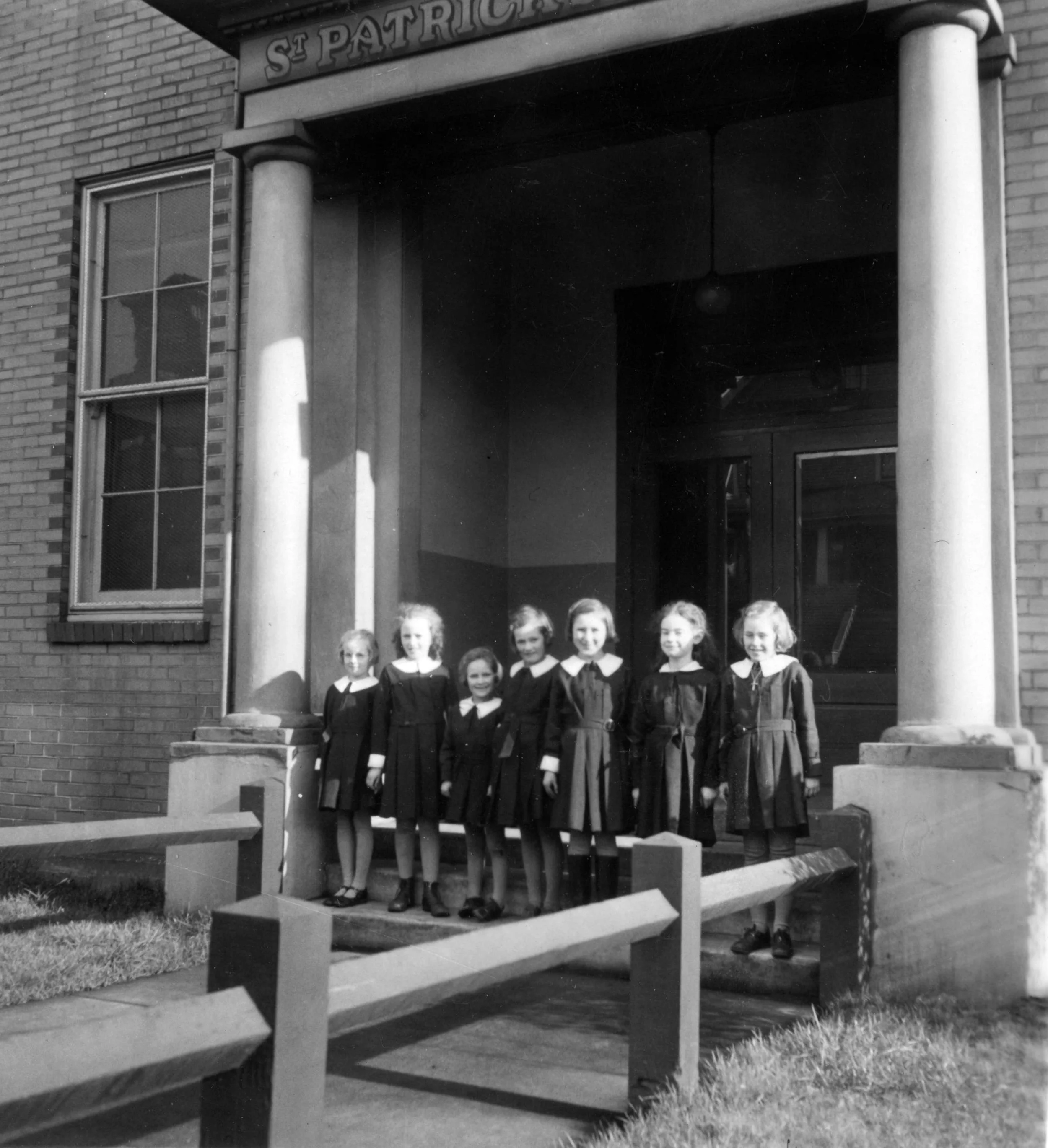 Black and white photograph of seven young girls standing in a row on the steps of a church building. The girls are dressed in dark dresses with white collars, and the sign above reads 'St. Patrick's.'