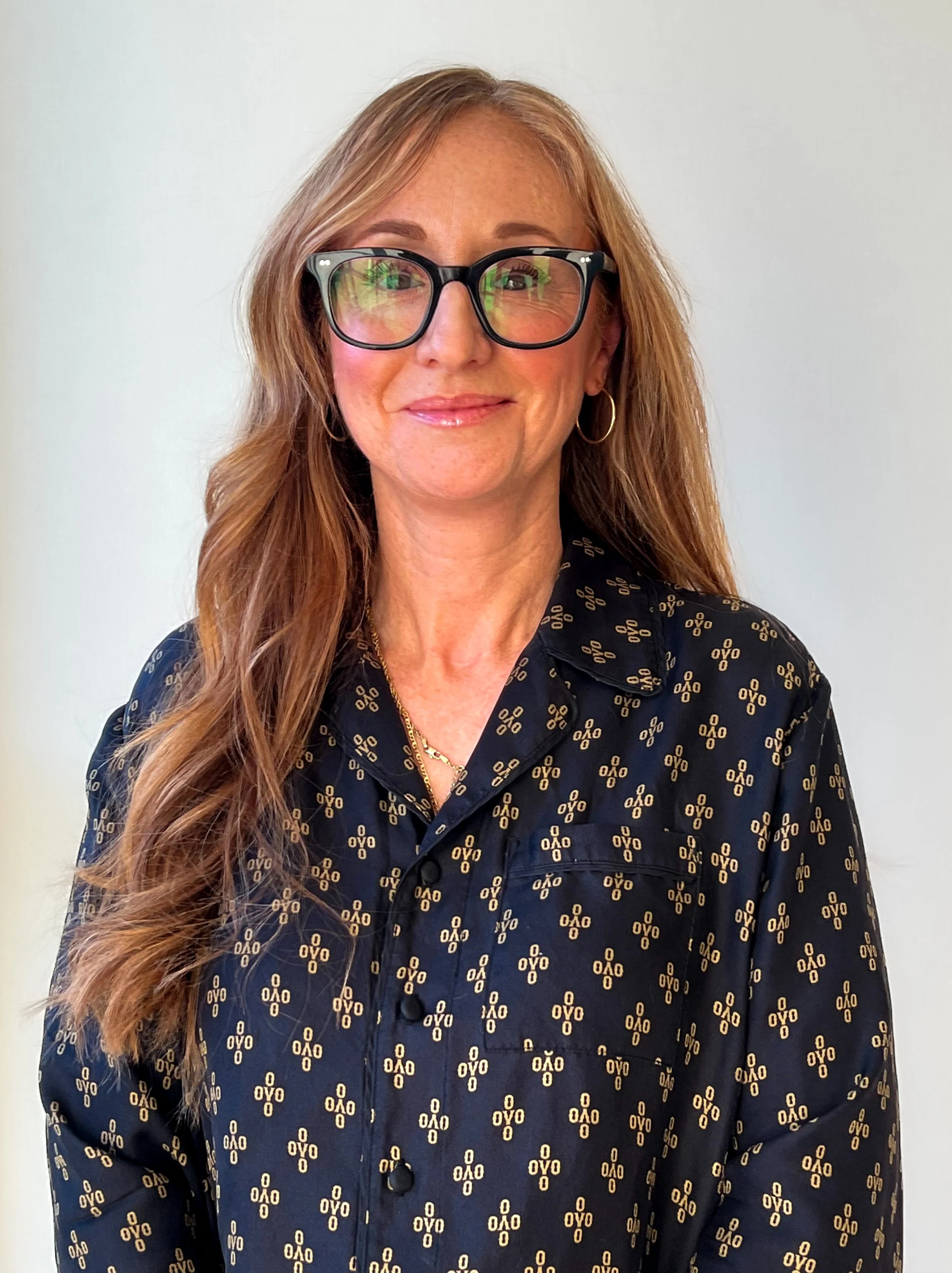 A woman with long, wavy, light brown hair wearing large black glasses, gold hoop earrings, a gold necklace, and a black patterned blouse with yellow design, smiling in front of a plain white background.