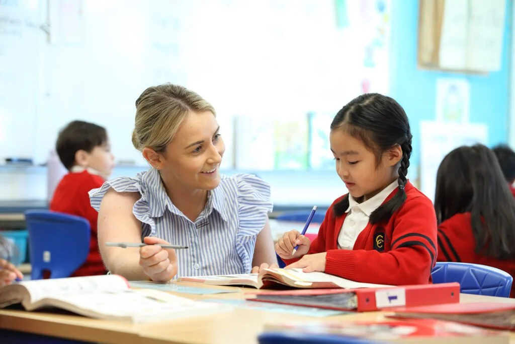 A female teacher smiling while assisting a young girl from an Asian background with braided hair, in a classroom setting with other students in the background.