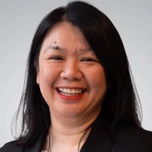 A woman smiling with dark hair and wearing a black blazer, facing the camera against a light gray background.