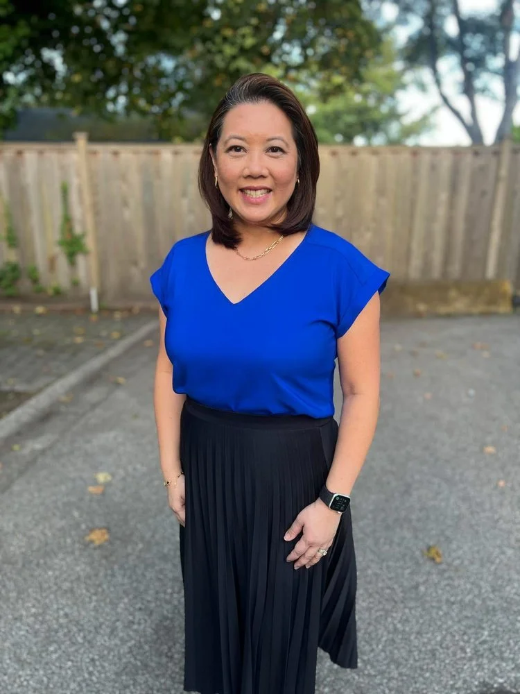 A woman with shoulder-length dark hair, wearing a blue top, black pleated skirt, gold necklace, bracelet, and an Apple Watch, standing outdoors on a paved surface near a wooden fence and trees.