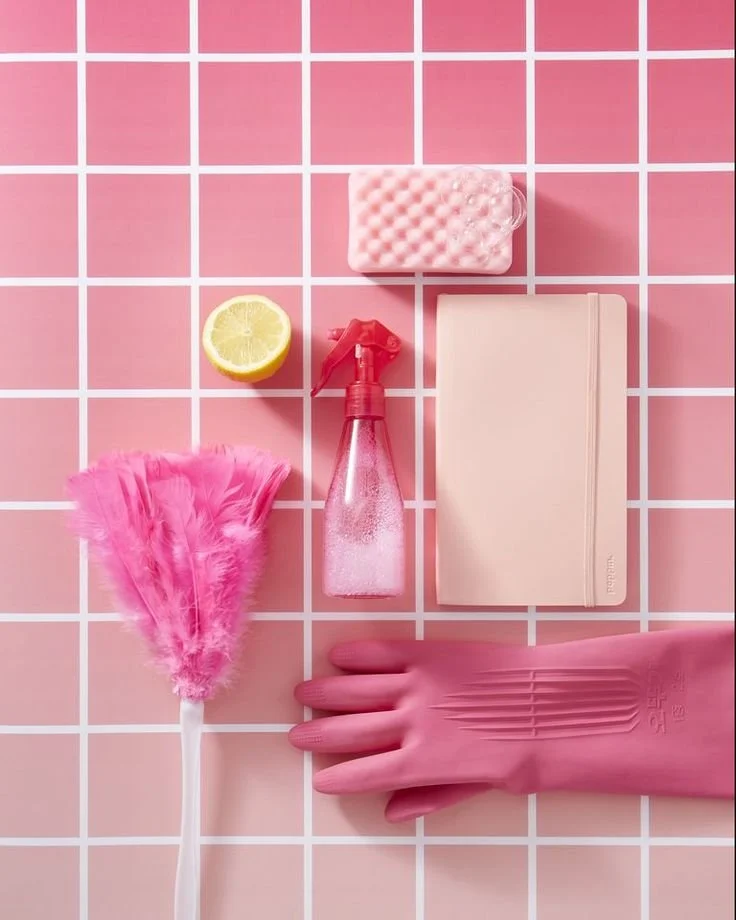 Cleaning supplies and a lemon slice arranged on pink tiled surface. Items include pink rubber gloves, pink scrub brush, spray bottle, bar soap, small notebook, and lemon half.