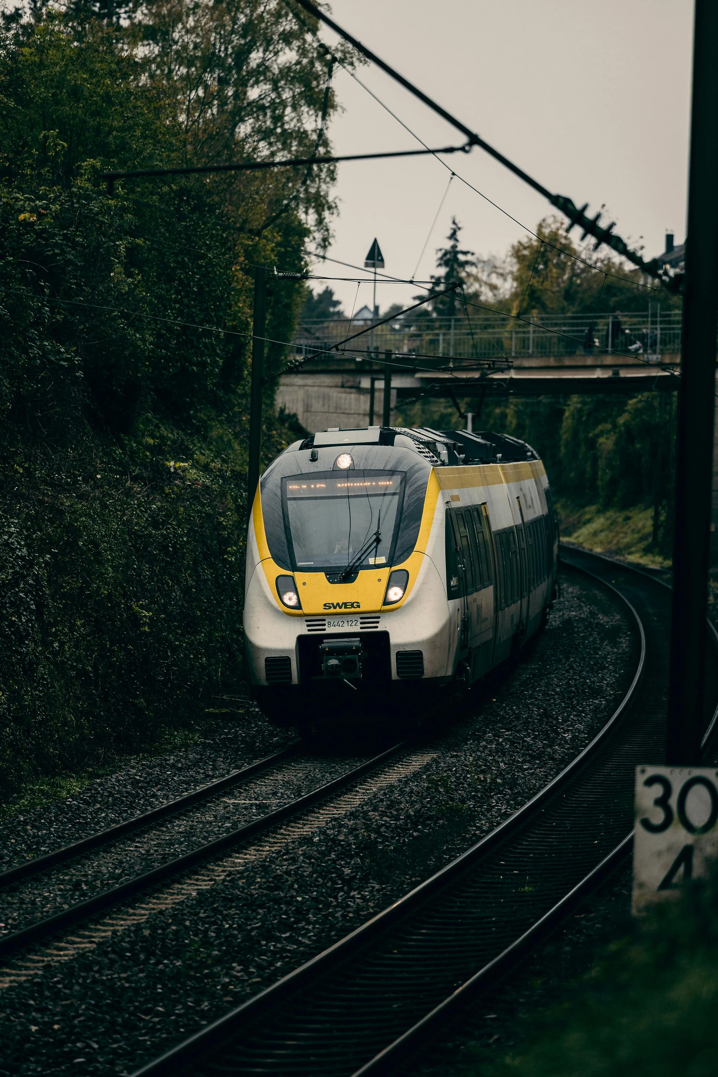 A modern yellow and gray passenger train traveling on tracks through greenery, with a bridge and trees in the background.