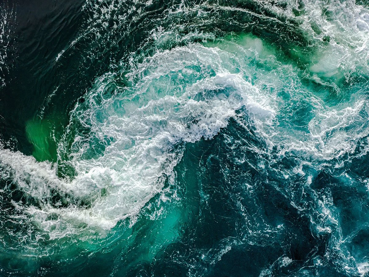 An aerial view of turbulent ocean water with swirling currents and white foam.