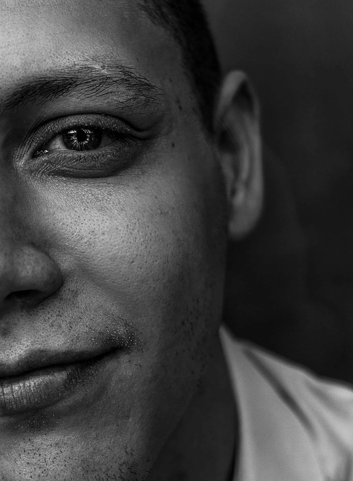 Close-up black and white photo of a smiling person's face, focusing on the left eye, cheek, and nose, with detailed skin textures and a blurred background.