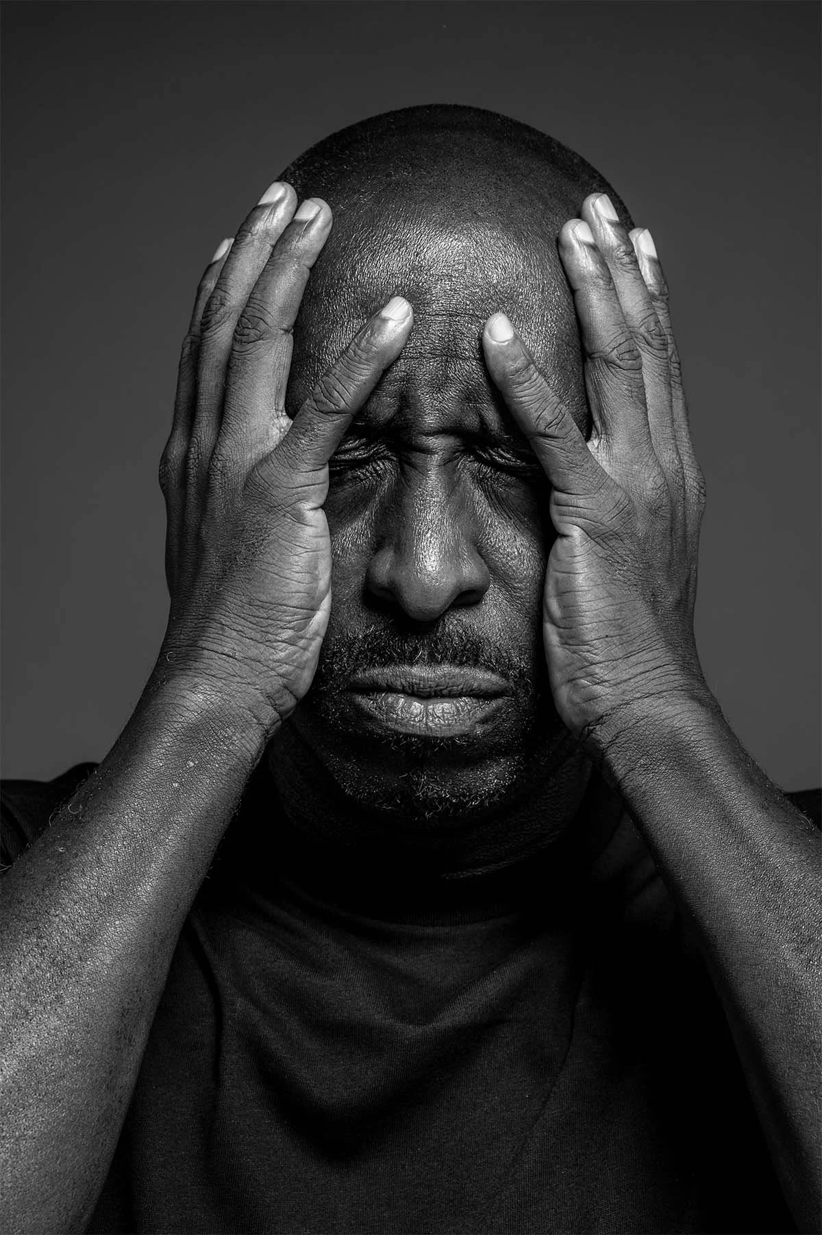 Black-and-white close-up of a man with his hands pressed to his temples, eyes closed and brow tense, conveying stress or emotional overwhelm.