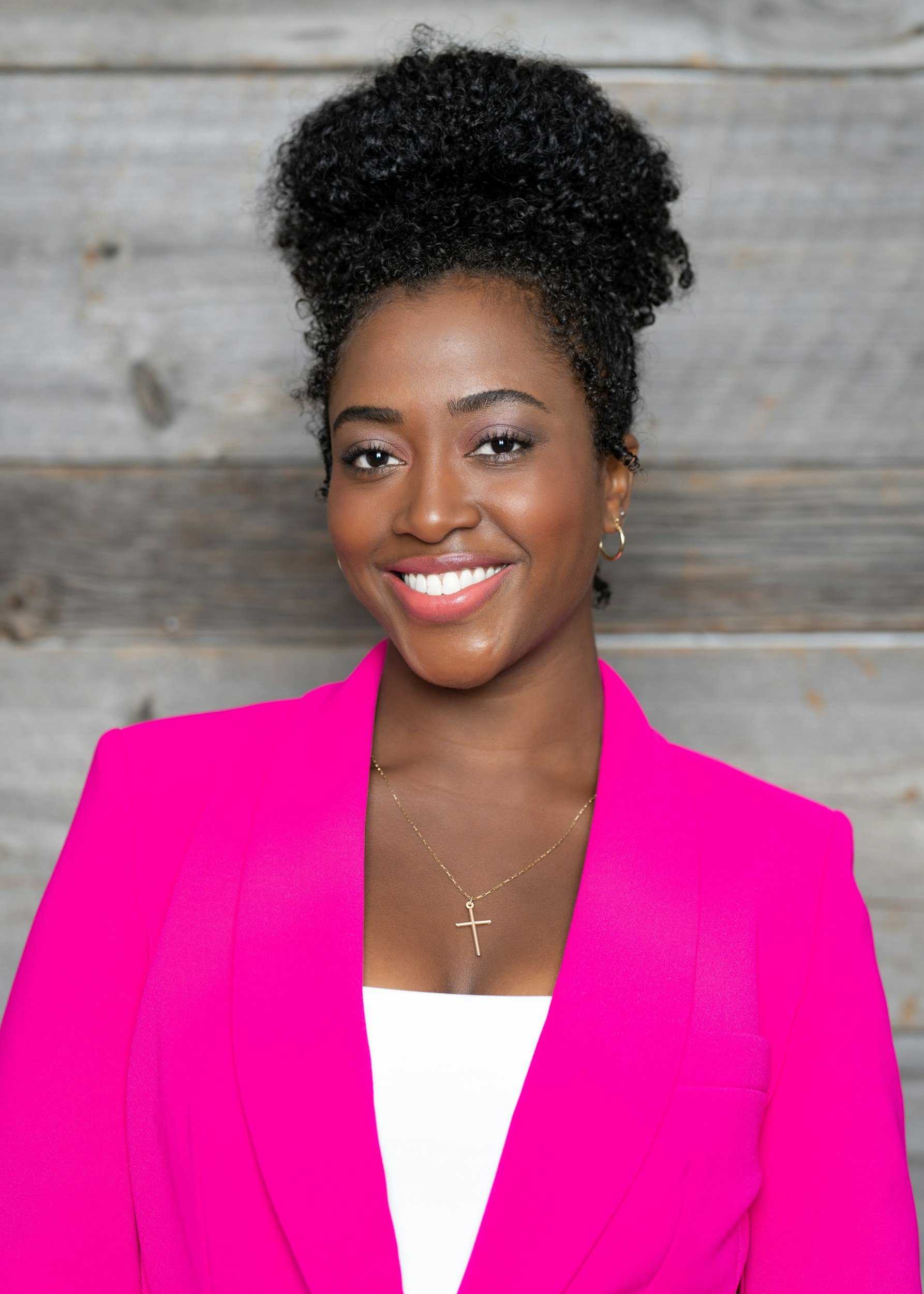 A smiling woman with dark curly hair styled in an updo, wearing a bright pink blazer, a white top, gold hoop earrings, and a gold cross necklace, standing in front of a rustic wooden background.