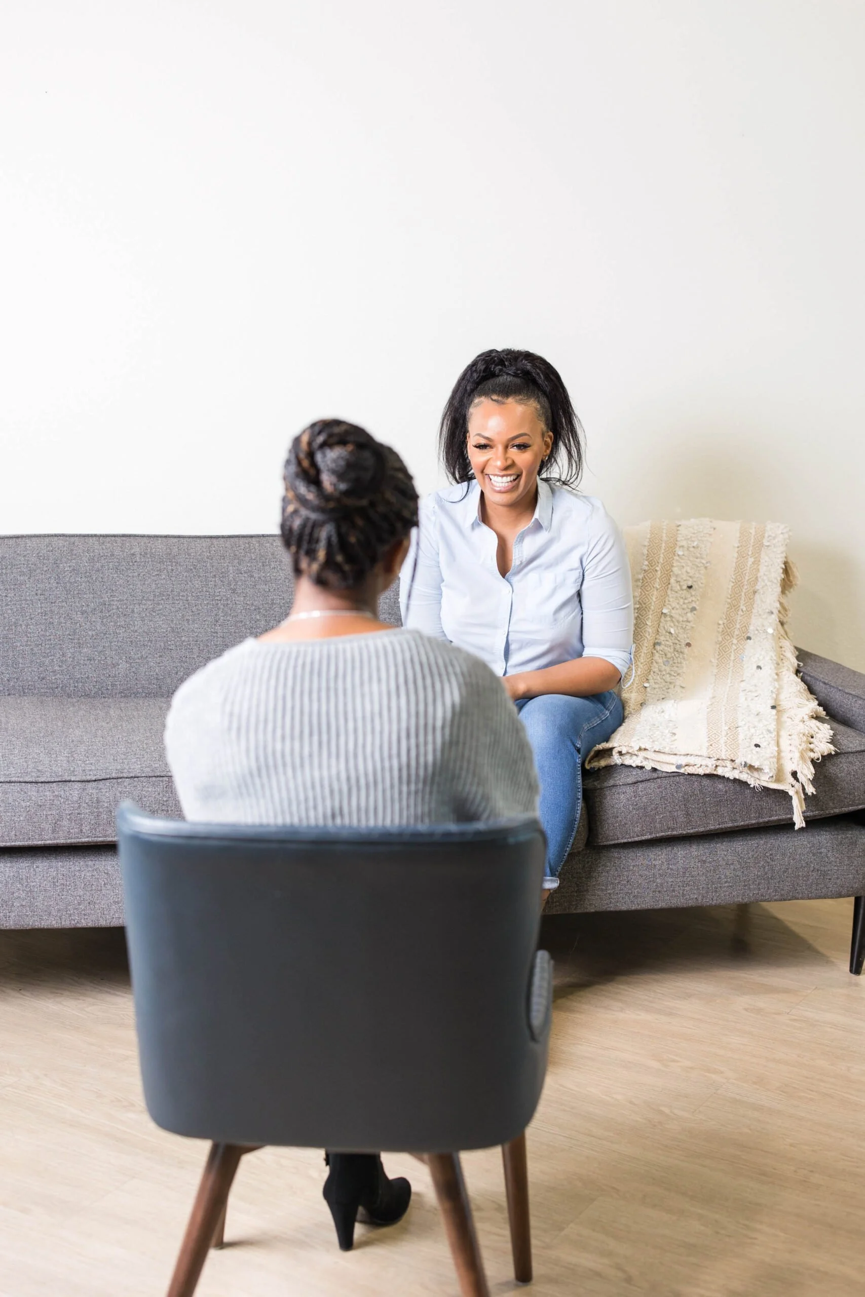 Two women sitting on a gray sofa, having a conversation. One woman with dark curly hair in a ponytail and the other with her back facing the camera, wearing a light sweater and black boots, in a cozy room with light wood flooring, a black chair, and a blanket on the sofa.