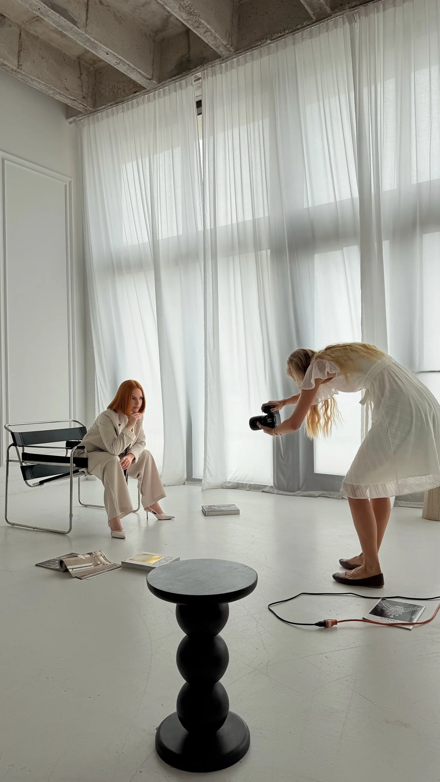 Female Photographer in white dress photprgpahing a red headed woman sitting in a chair wearing a business suit.