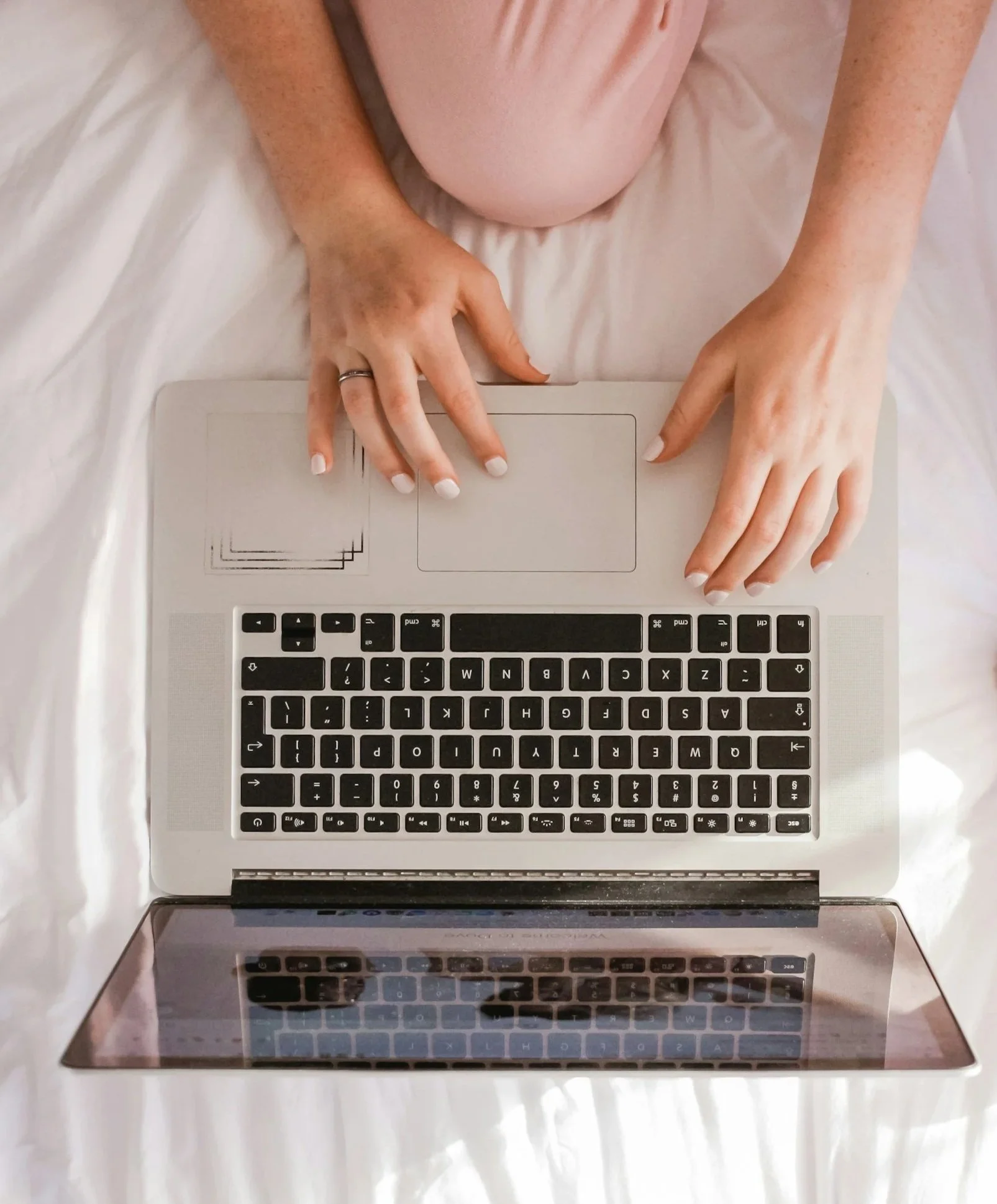Top-down view of a person with painted nails using a silver laptop on a white bed.