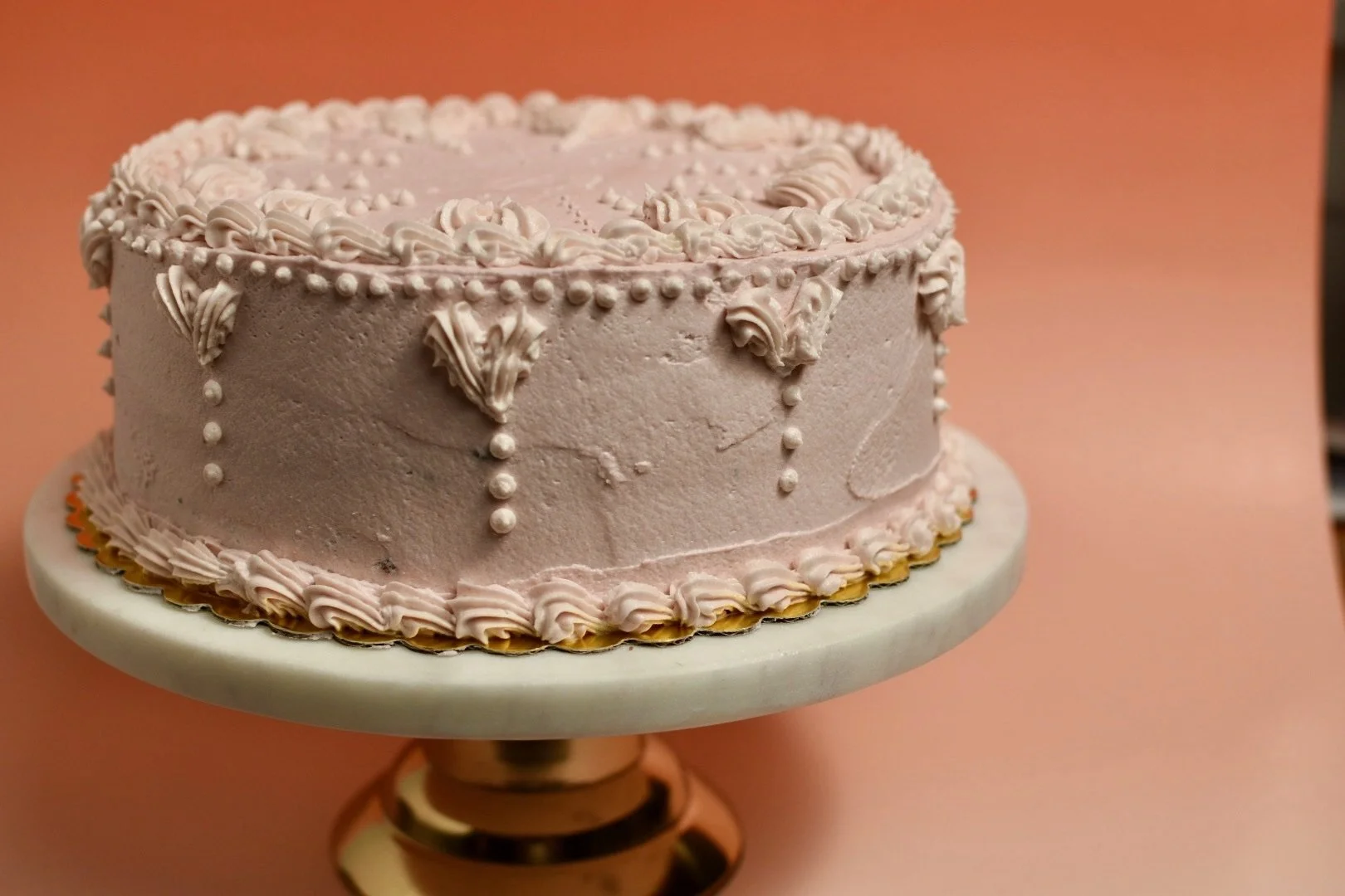 A strawberry frosted plant-based, vegan birthday cake decorated with piped icing and edible pearls, placed on a white cake stand with a gold base, against a peach-colored background.