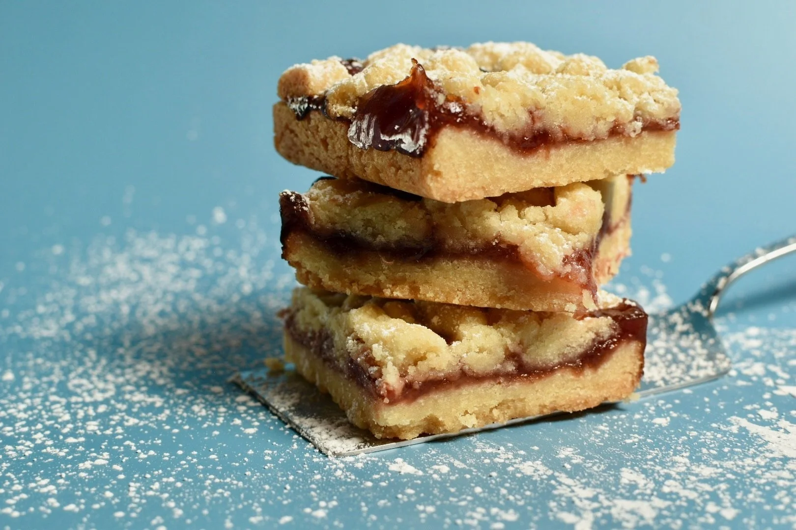 Three stacked slices of vegan raspberry crumble bar on a blue background, dusted with powdered sugar.