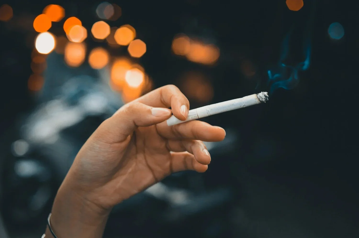 A hand holding a lit cigarette with smoke rising from it, against a blurred background with warm and cool lights.