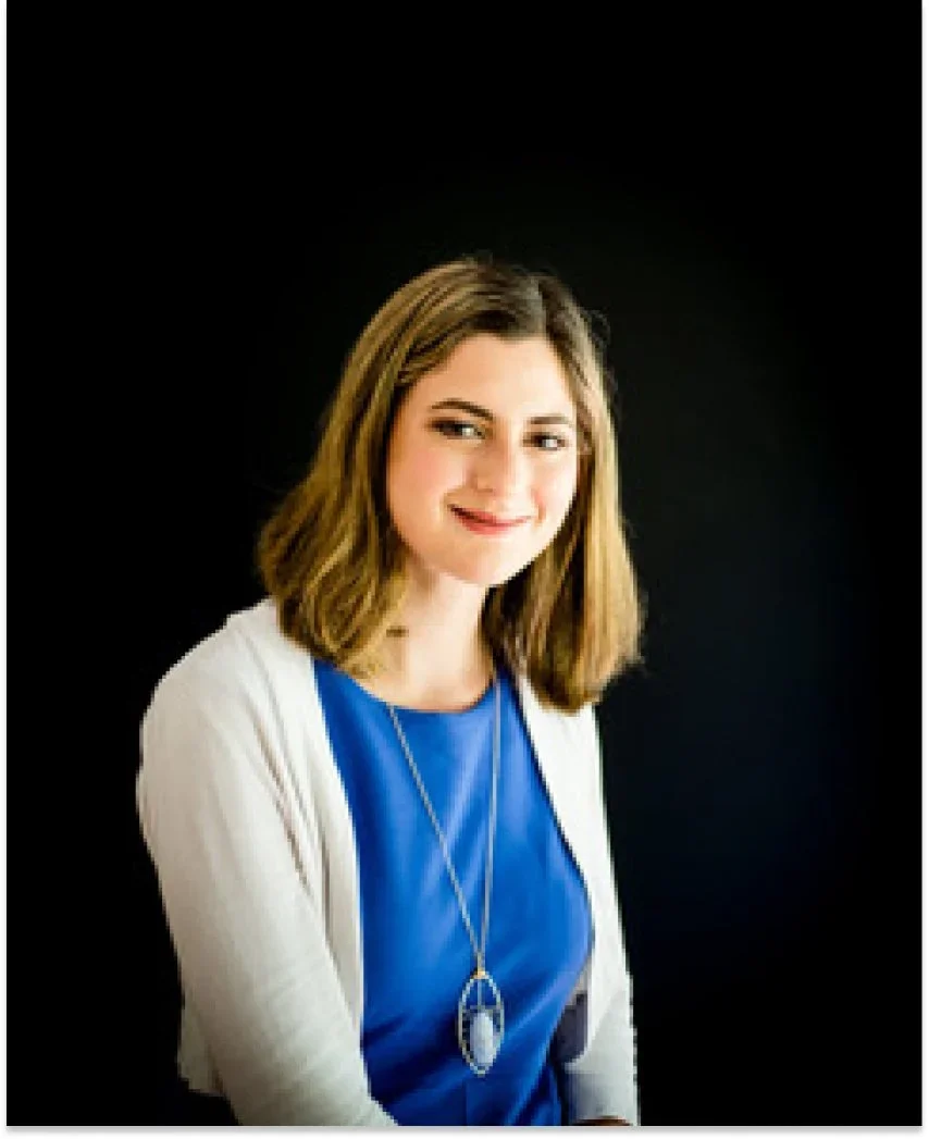 A woman with shoulder-length brown hair and a friendly smile, wearing a blue top, white cardigan, and a long necklace, posing against a black background.