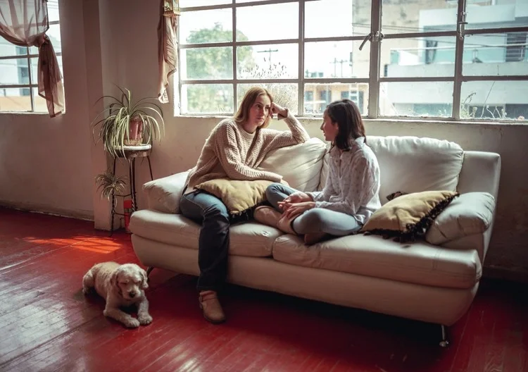 Two women sit on a white couch in a sunlit room with large windows, talking. A small puppy is lying on the wooden floor nearby.