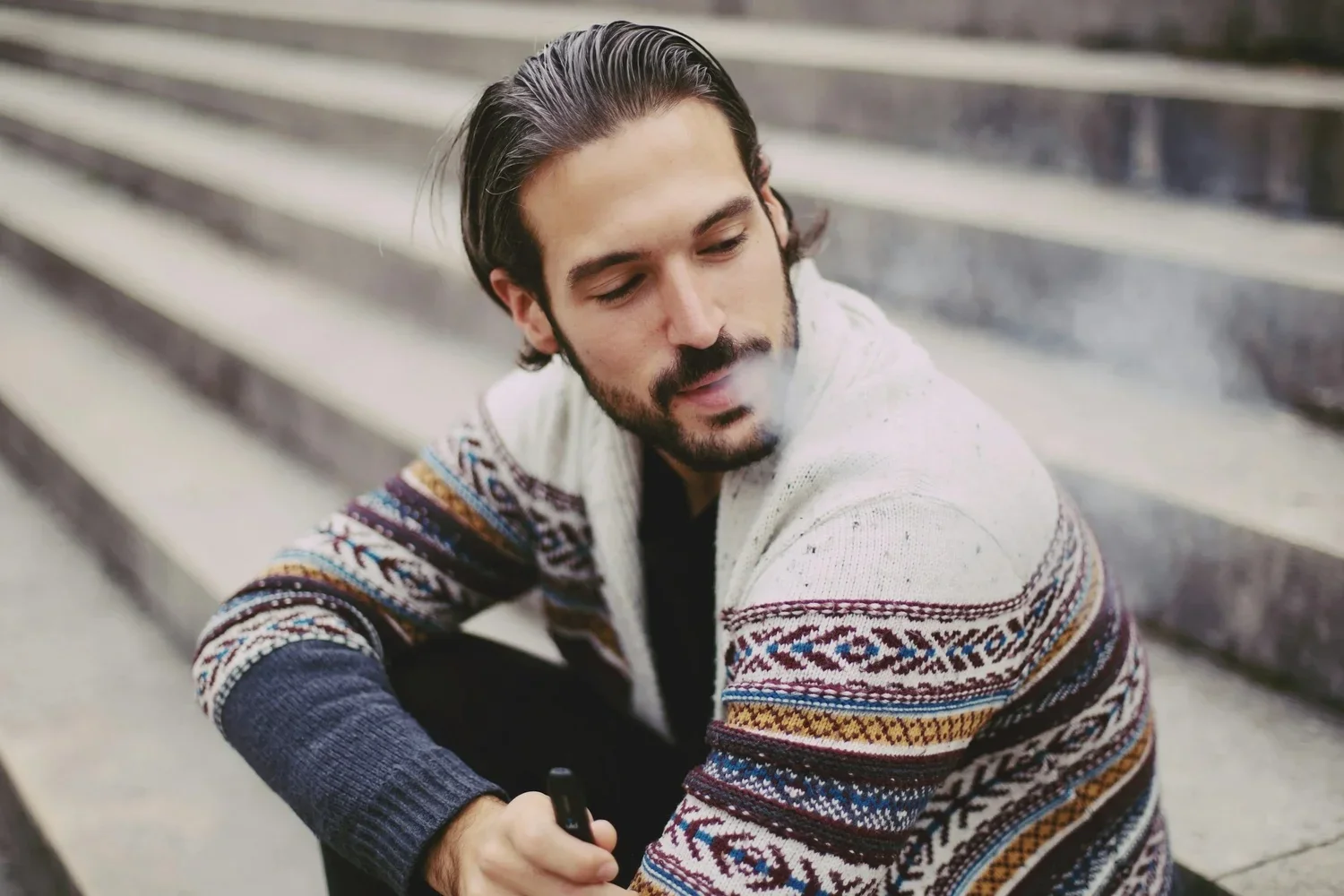 A man with dark hair and a beard sitting on outdoor steps, wearing a patterned sweater, exhaling smoke while holding a vape or cigarette.