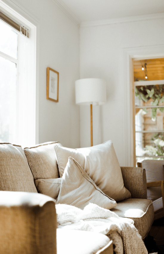 Cozy living room with beige sofa, cushions, and a blanket, illuminated by natural light from a window, with a white lamp and framed artwork on the wall.