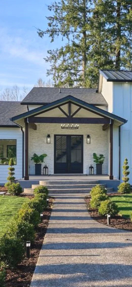 Front porch of a modern house with black double doors, two large planters with green plants, and a gabled roof. A gravel walkway with small bushes leads to the entrance, with neatly maintained green lawn on both sides and solar pathway lights. Tall trees and a partly cloudy sky are in the background.