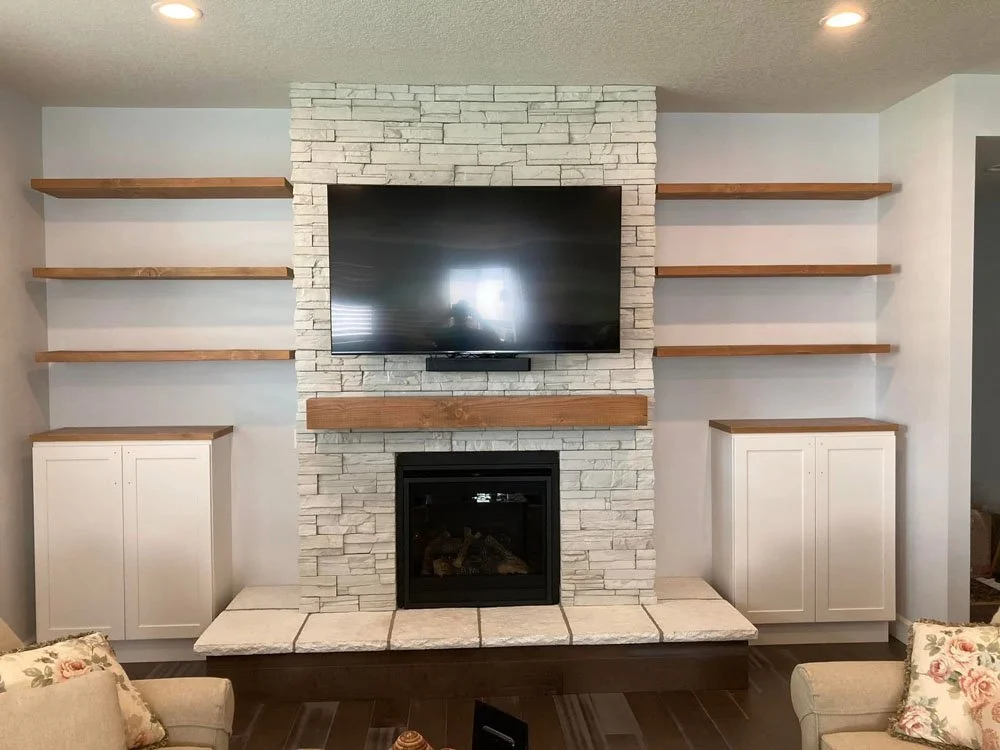 Living room with white stacked stone fireplace, wall-mounted flat-screen TV, wooden shelves, white cabinets, cushioned chairs, and floral pillows.