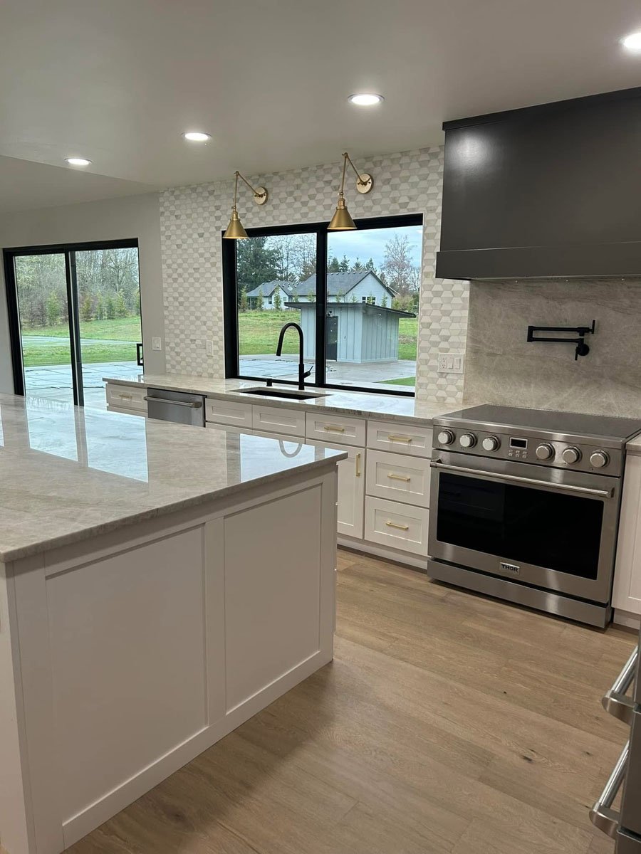 Modern kitchen with white cabinets, marble countertops, a stainless steel oven, black faucet, and large windows showing a backyard with grass and trees.