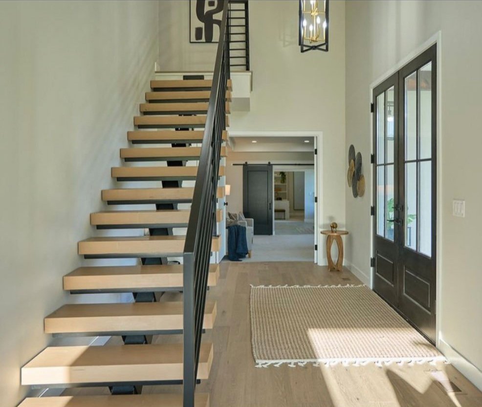 Entryway with a wooden staircase, glass-paneled black front door, beige walls, a rectangular rug, and a view into a living space with a black sliding barn door.