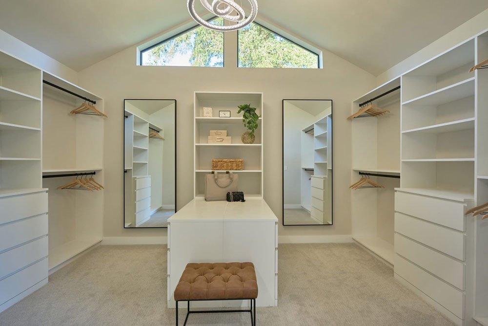 Modern walk-in closet with white shelving units, mirrors, and a small brown tufted bench, a window near the ceiling, and a central white dresser.