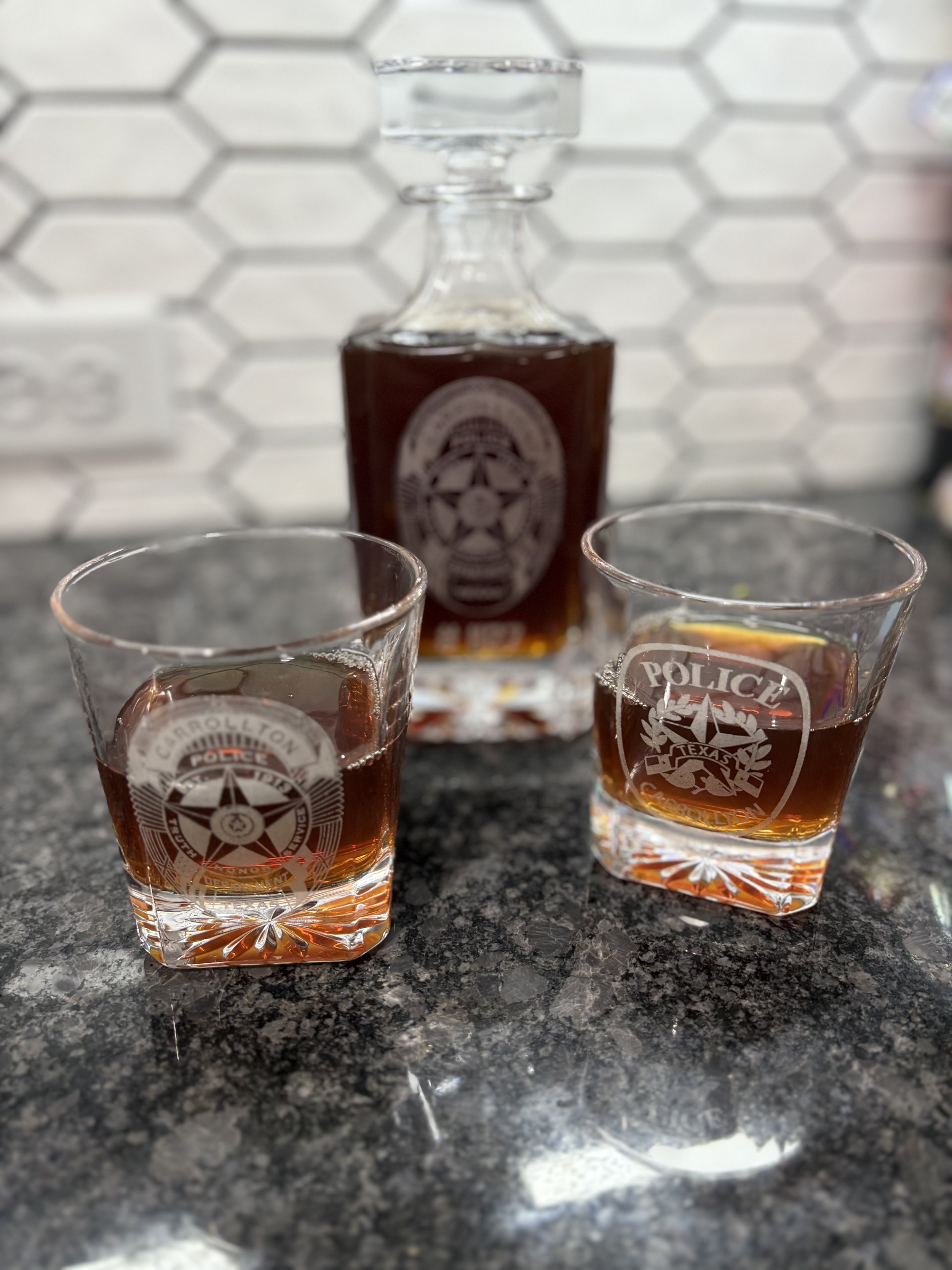 A carrollton police whiskey decanter with two glasses of whiskey in front of it on a dark granite countertop with a white hexagonal tile backsplash in the background.