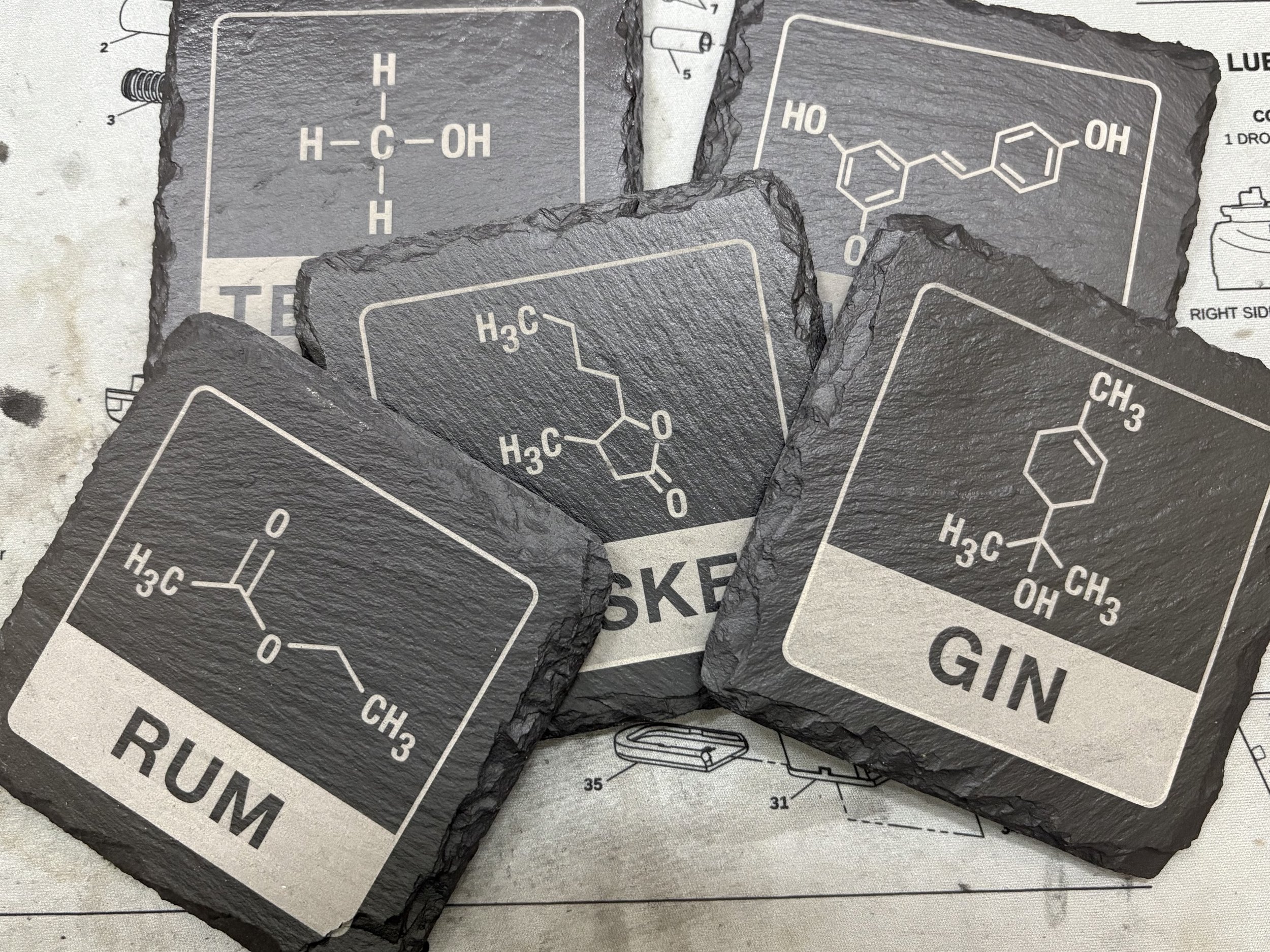 Set of four black slate coasters featuring chemical structures and names of alcoholic beverages: Rum, Gin, Tequila, and an unidentified beverage.
