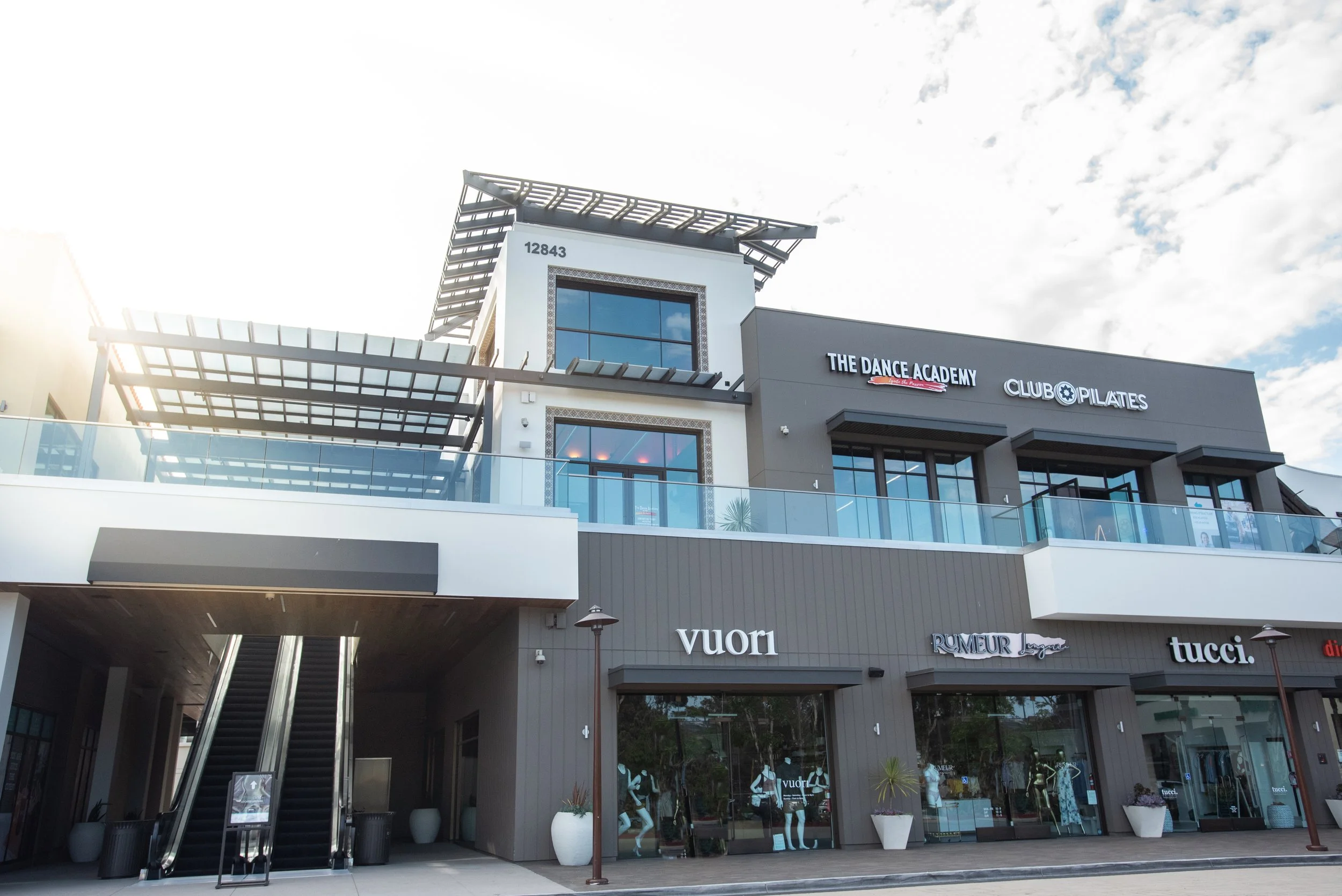 A modern shopping center with multiple store signs including Vuori, The Dance Academy, Club Pilates, Rumeur Linge, and Tucci. The building has large windows, a glass staircase, and tiered architecture with open space on top, set against a partly cloudy sky.