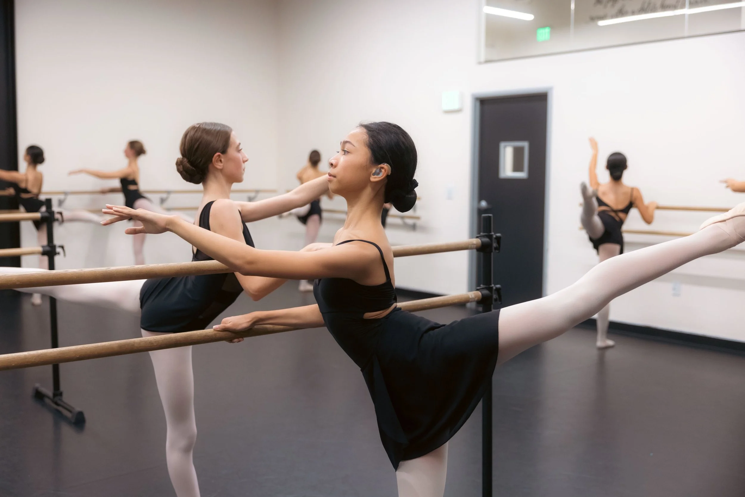 Ballet dancers in a dance studio practicing at the ballet barre, with some stretching and performing ballet poses.