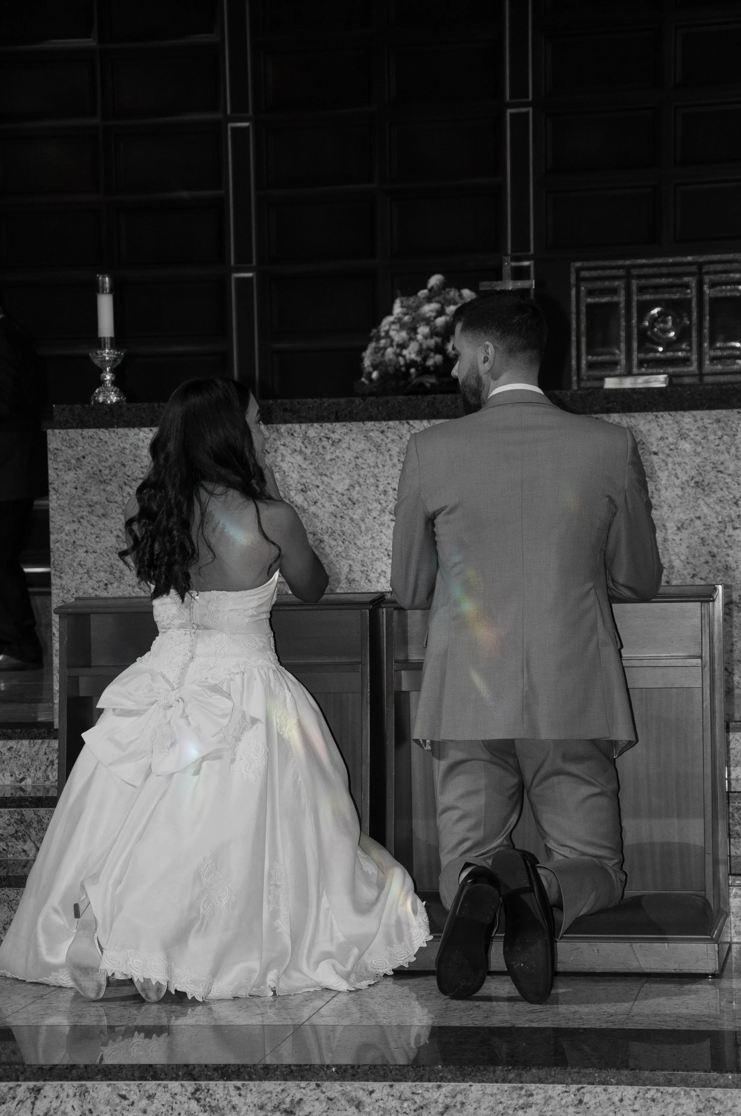 Bride and groom kneeling at altar, wearing wedding dress and suit, in black and white photo.