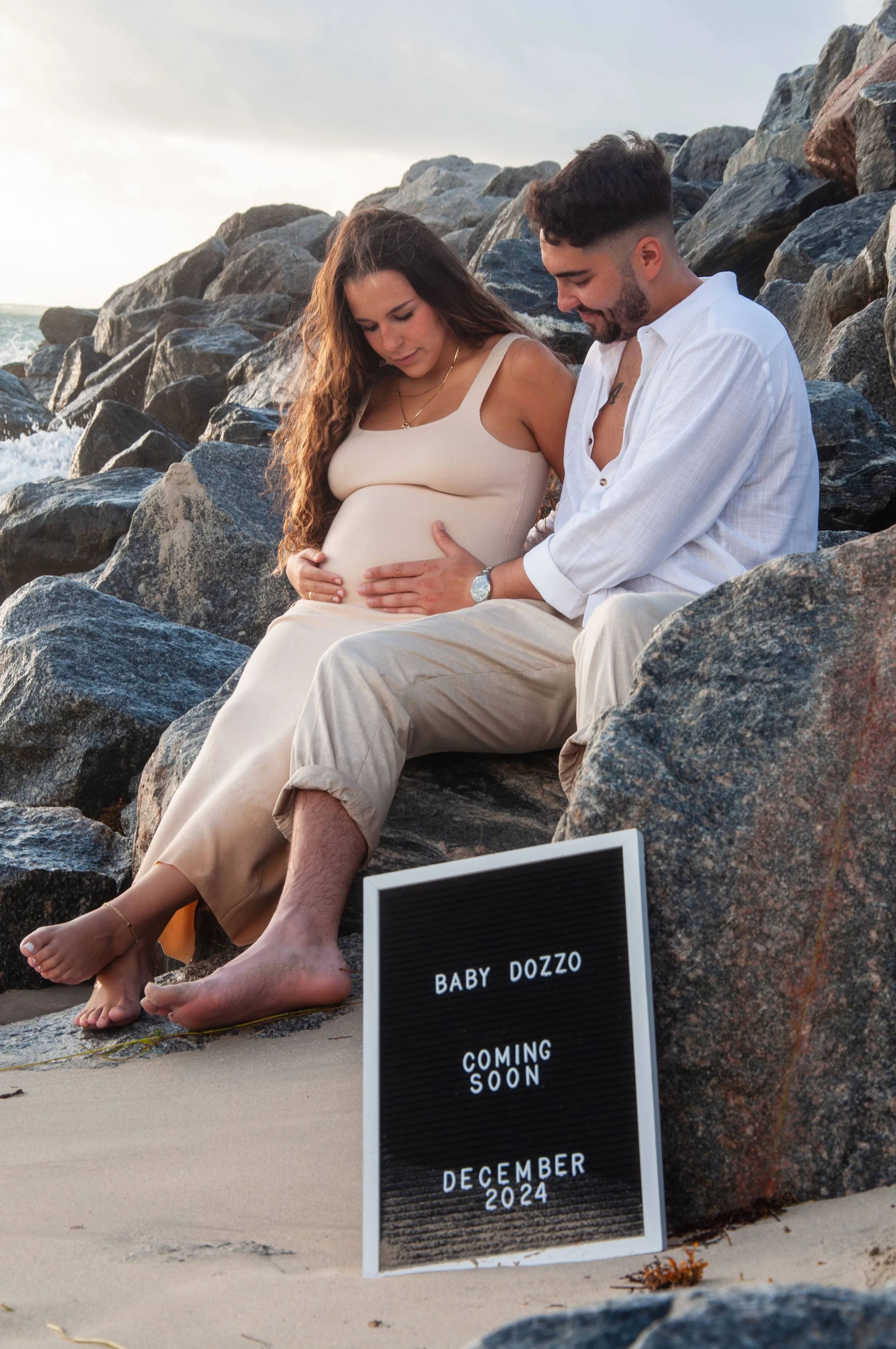 Pregnant woman and partner sitting on rocks by the ocean, holding her belly, with a sign saying 'Baby Dozzo Coming Soon December 2024.'