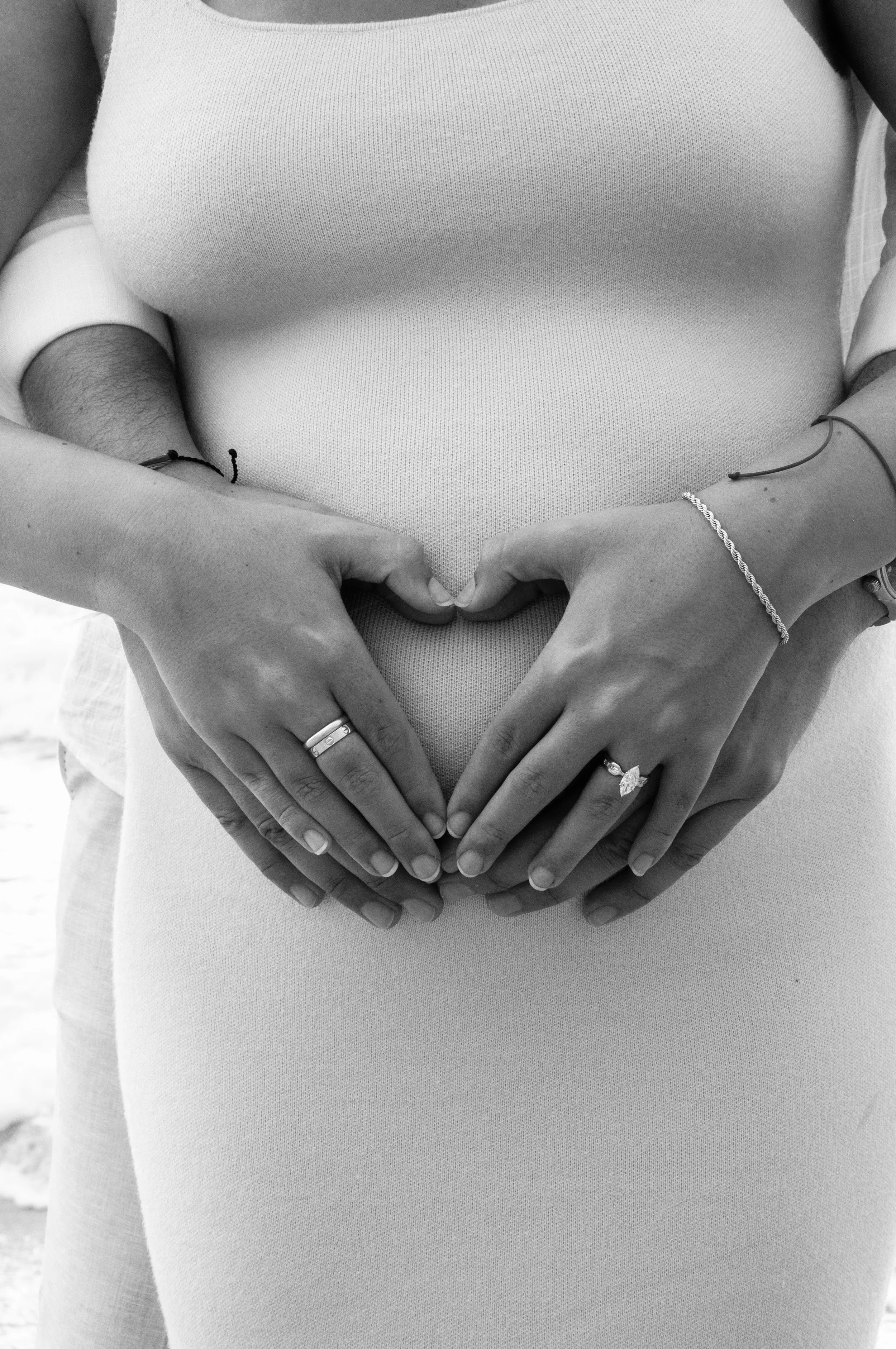 Close-up of a pregnant woman with a man's arms around her, both forming a heart shape over her belly with their hands.